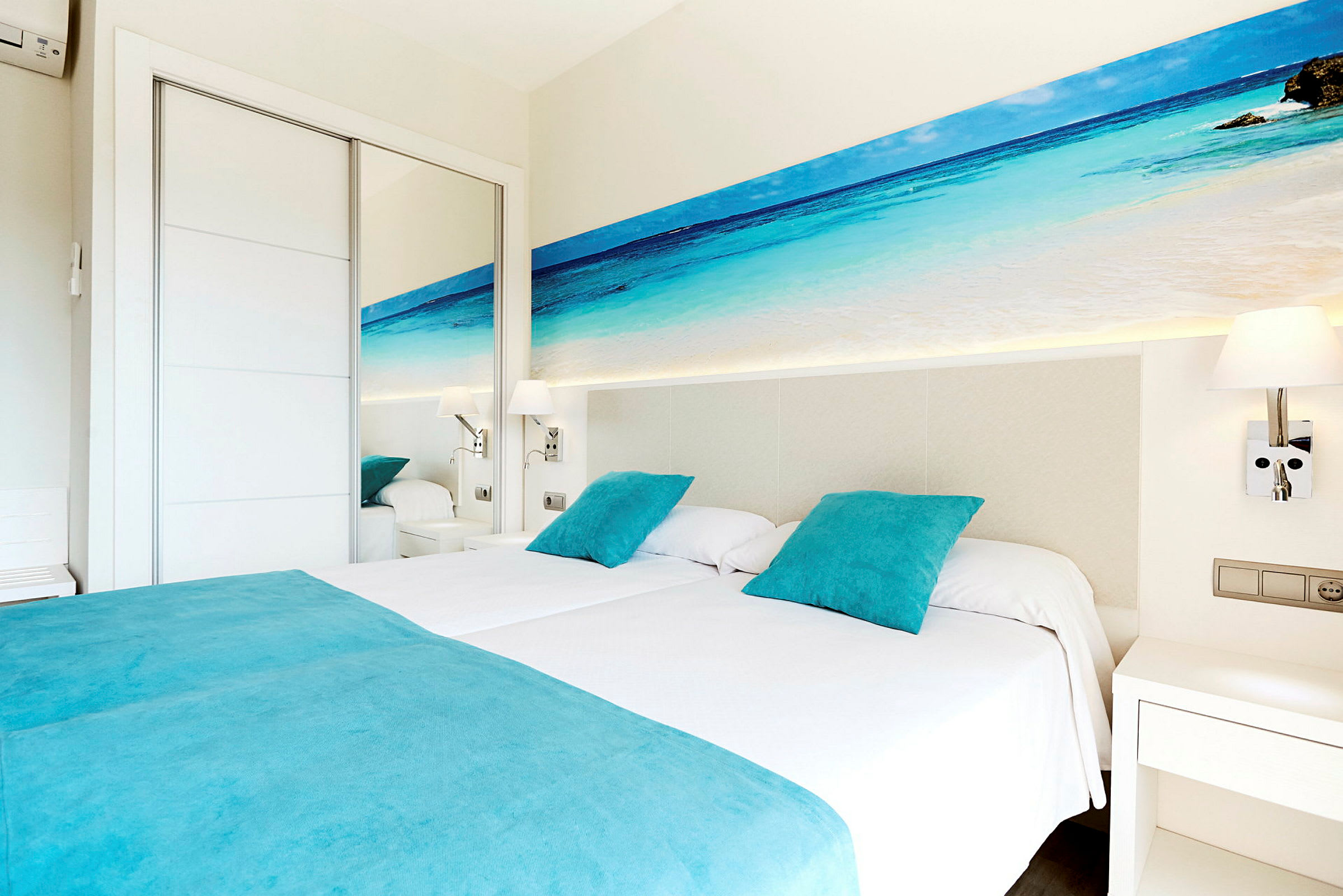 Ett hotellrum med två enkelsängar, bäddade med vita lakan och turkosa kuddar och överkast. På väggen ovanför sängarna finns en bild av en strand med hav. Rummet har vita möbler och en spegel. (AI-genererad text)