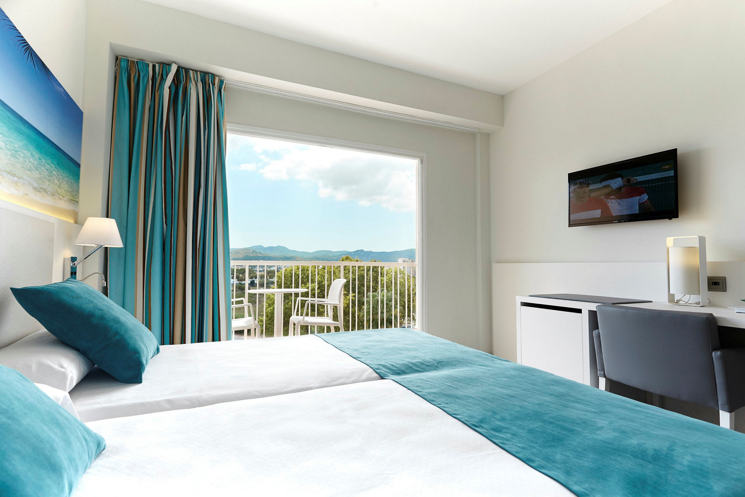 Ett hotellrum med två enkelsängar, en tavla med strandmotiv på väggen, stora fönster med utsikt över landskap, och en TV på väggen. (AI-genererad text)