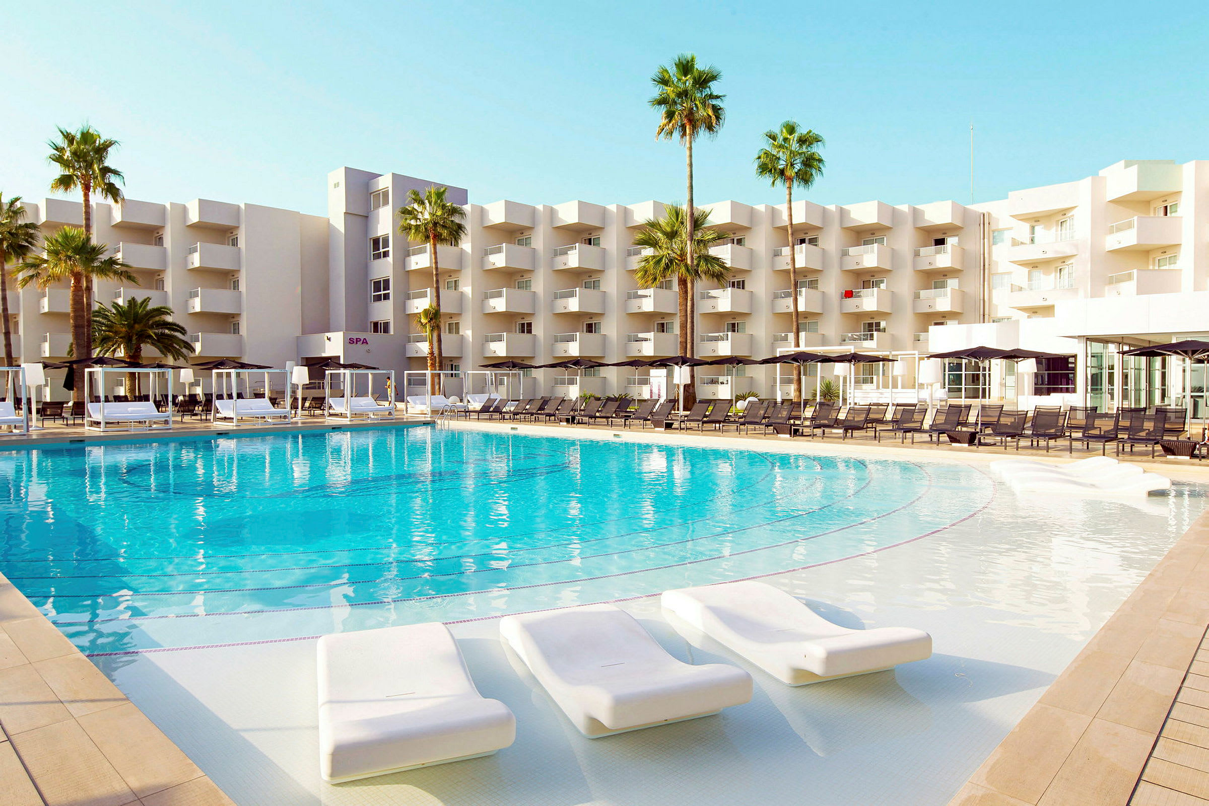 Stor simbassäng med omgivande solstolar framför en modern hotellbyggnad och palmer. (AI-genererad text)