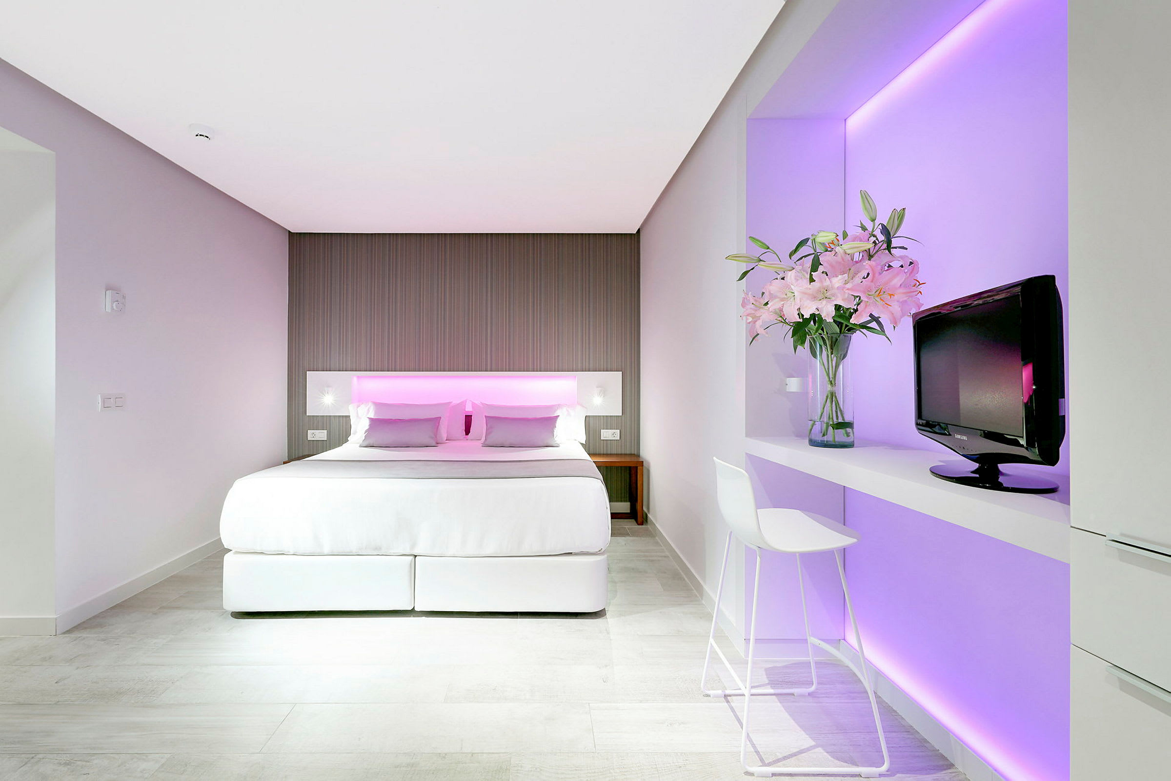 Hotellrum med dubbelsäng, nätt inredning och rosa belysning bakom sänggaveln. En TV och en vas med blommor står på en hylla bredvid. (AI-genererad text)
