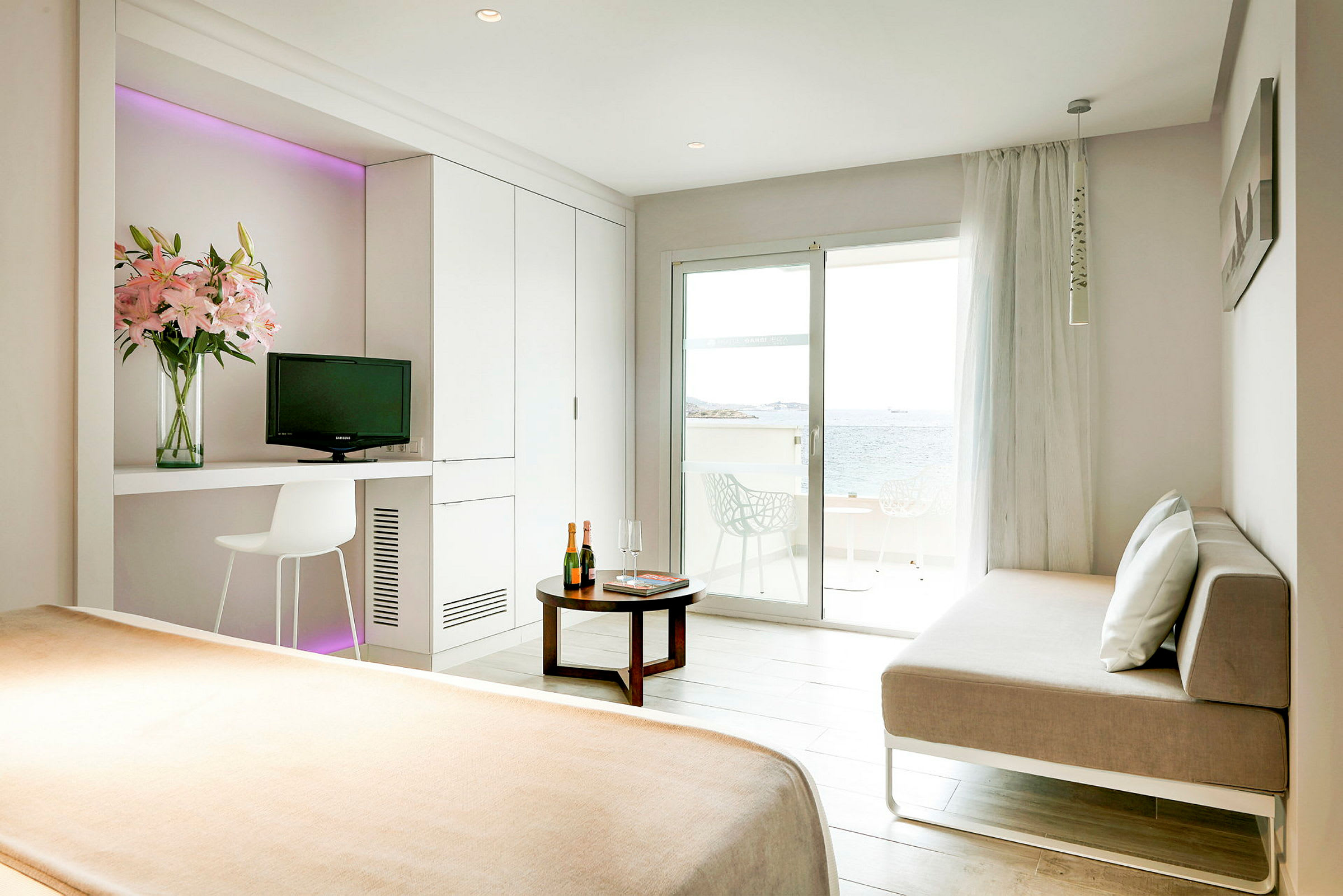 Ett ljust hotellrum med en säng, en soffa, ett bord och en balkong som vetter mot havet. (AI-genererad text)