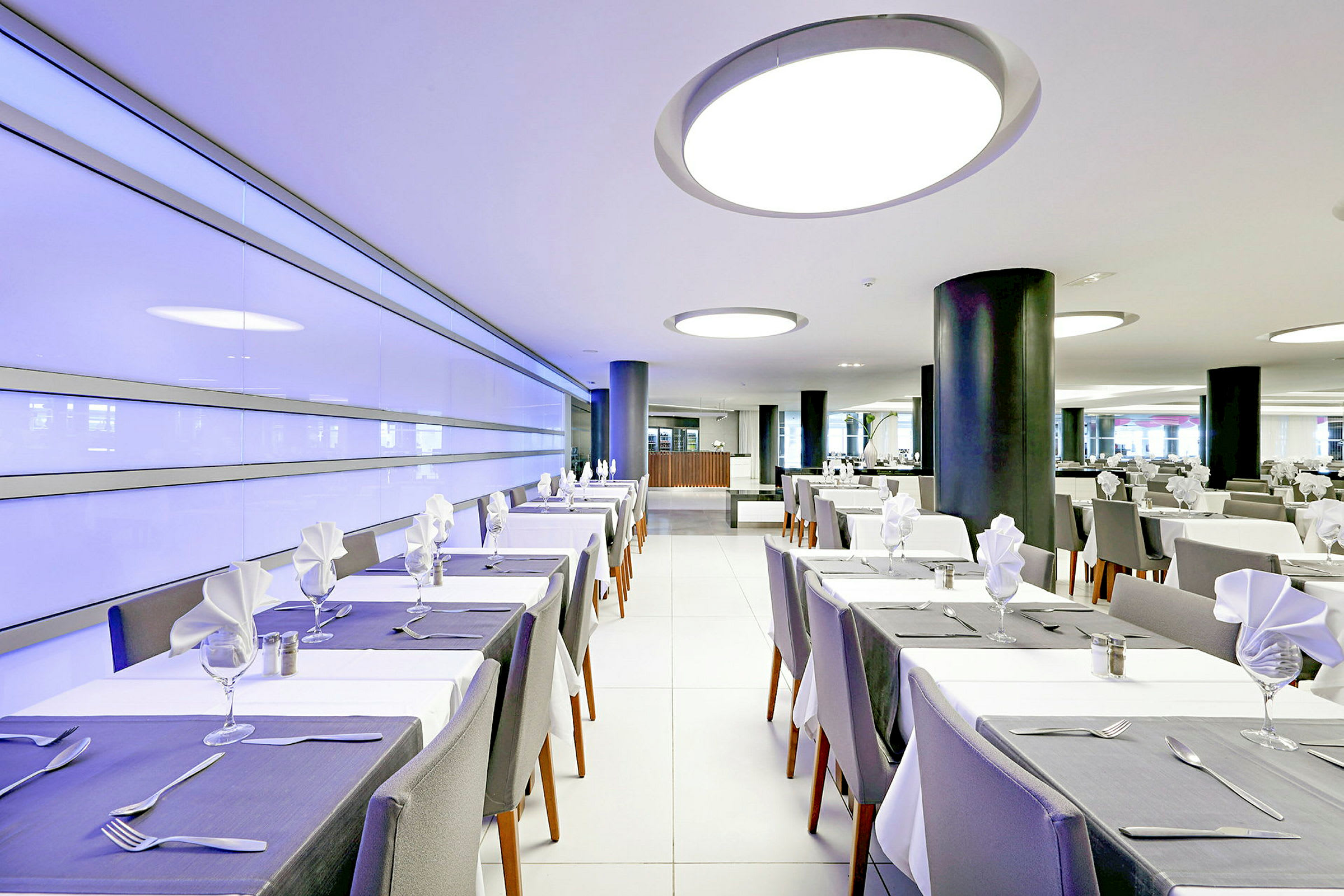 En modern restaurang med dukade bord och vita stolar. Inredningen inkluderar moderna lampor och en ljus atmosfär. (AI-genererad text)