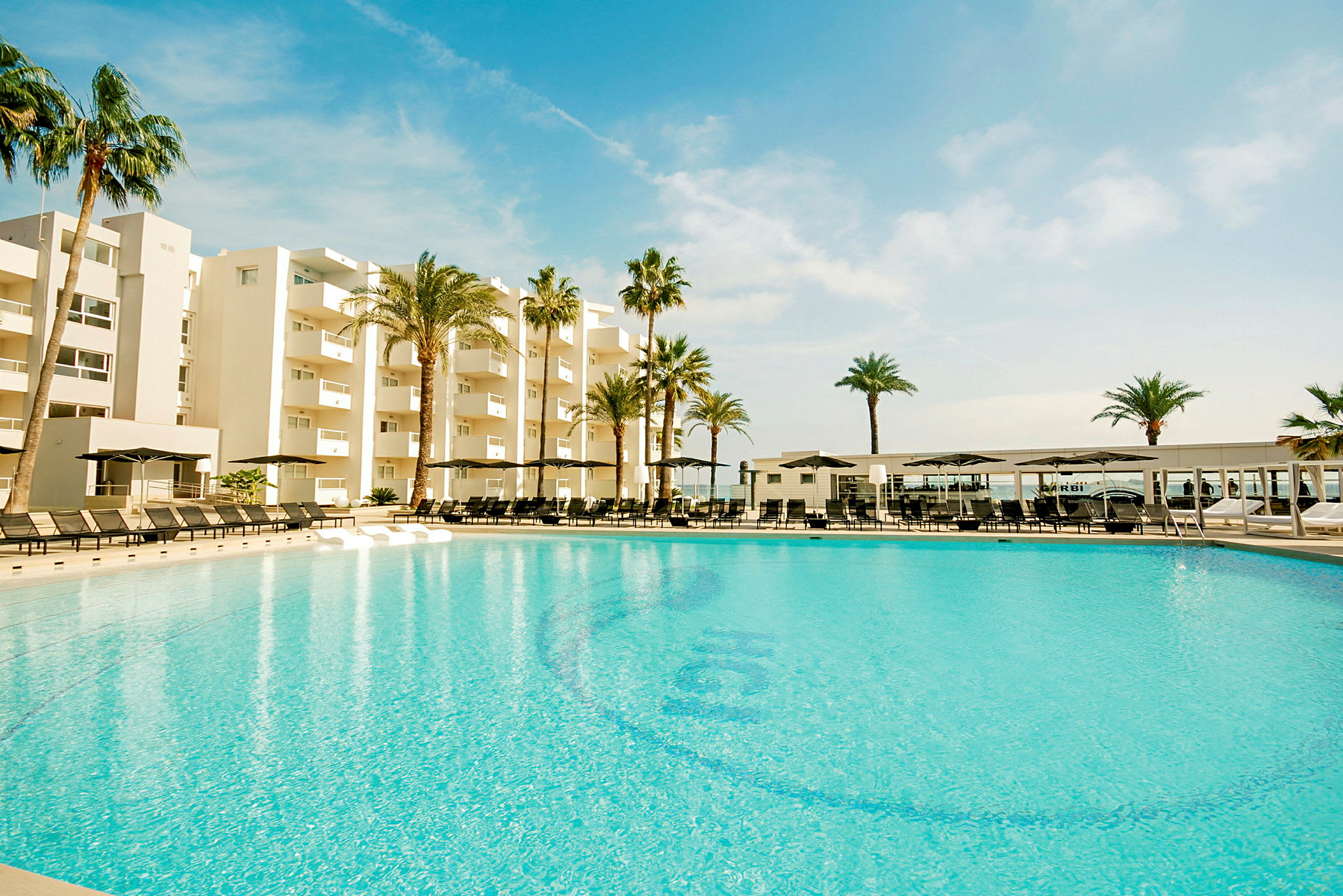 En stor pool framför ett modernt hotell omgiven av palmer under en blå himmel. (AI-genererad text)