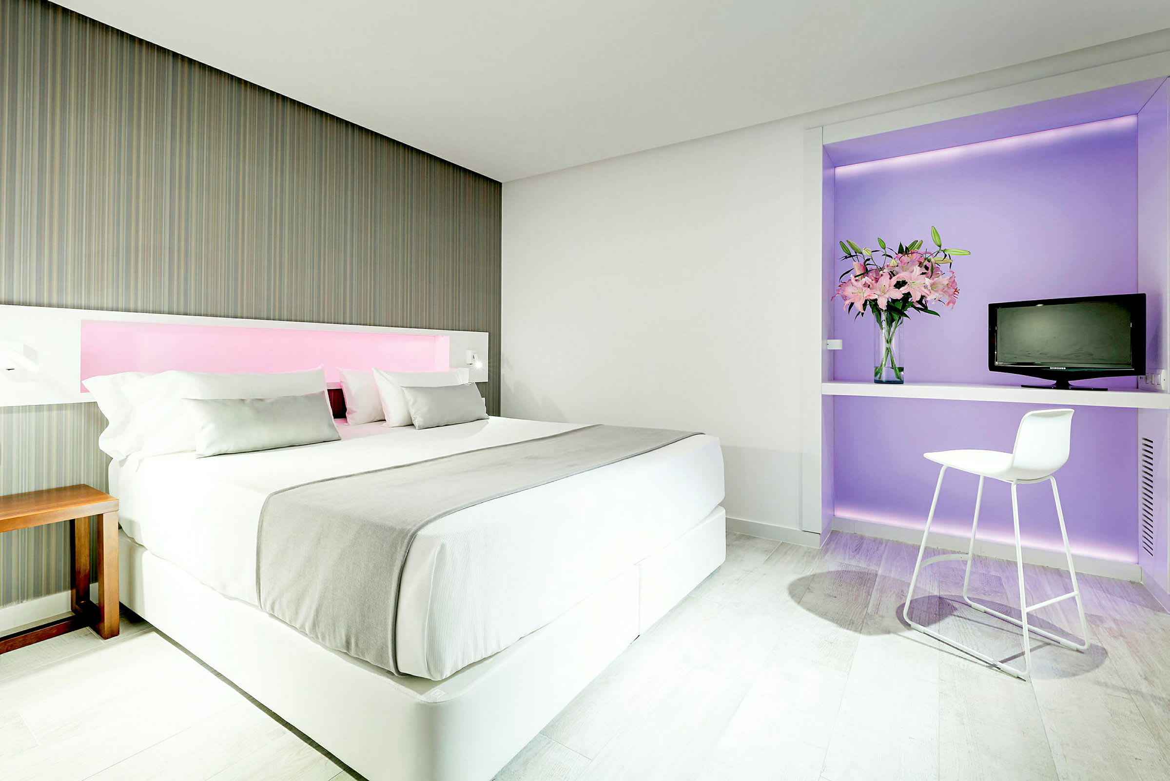 Ett modernt hotellrum med en stor säng, dekorerat i ljusa färger. Till höger finns ett skrivbord med en stol, en vas med blommor och en liten TV. (AI-genererad text)