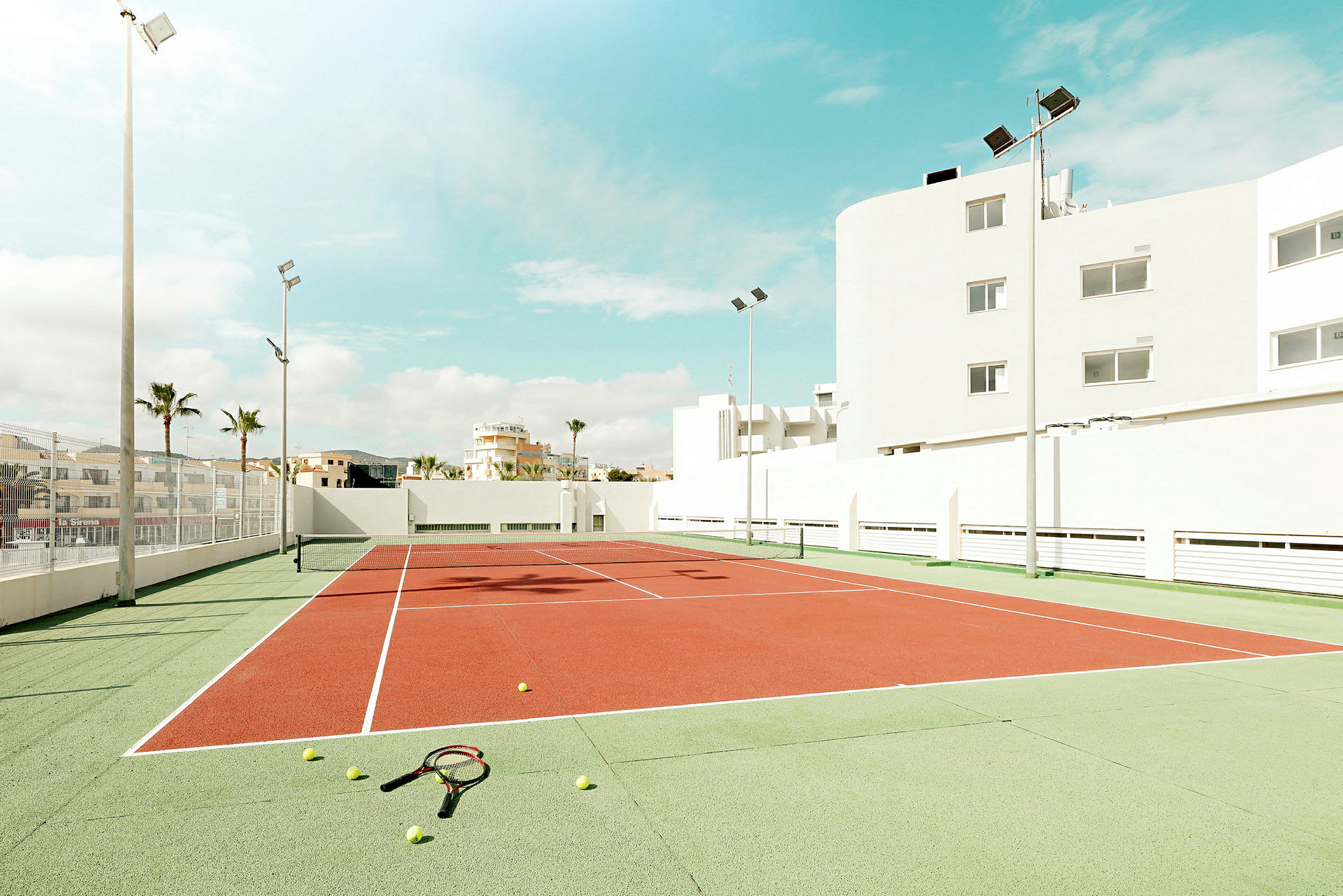 En tennisbana omgiven av byggnader under en solig himmel, med tennisbollar och racketar liggande på marken. (AI-genererad text)