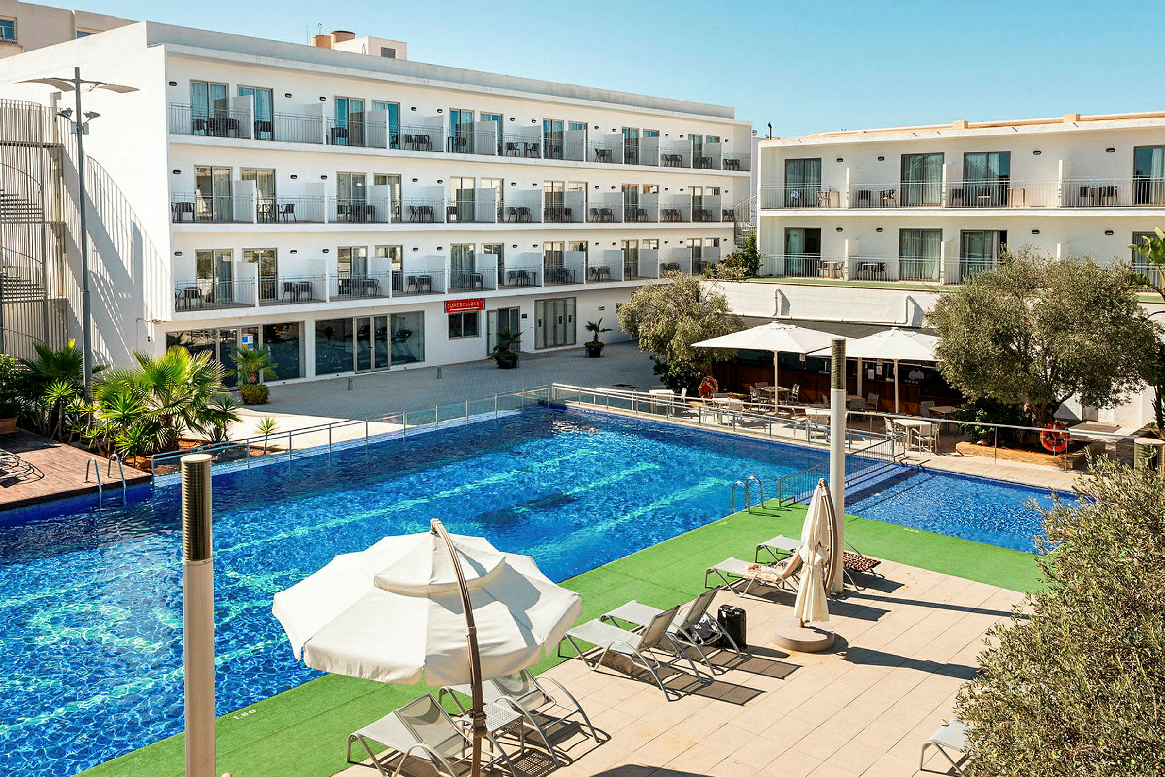 Bild av en hotellbyggnad med flera våningar. Framför byggnaden finns en rymlig pool och solstolar under parasoller. (AI-genererad text)