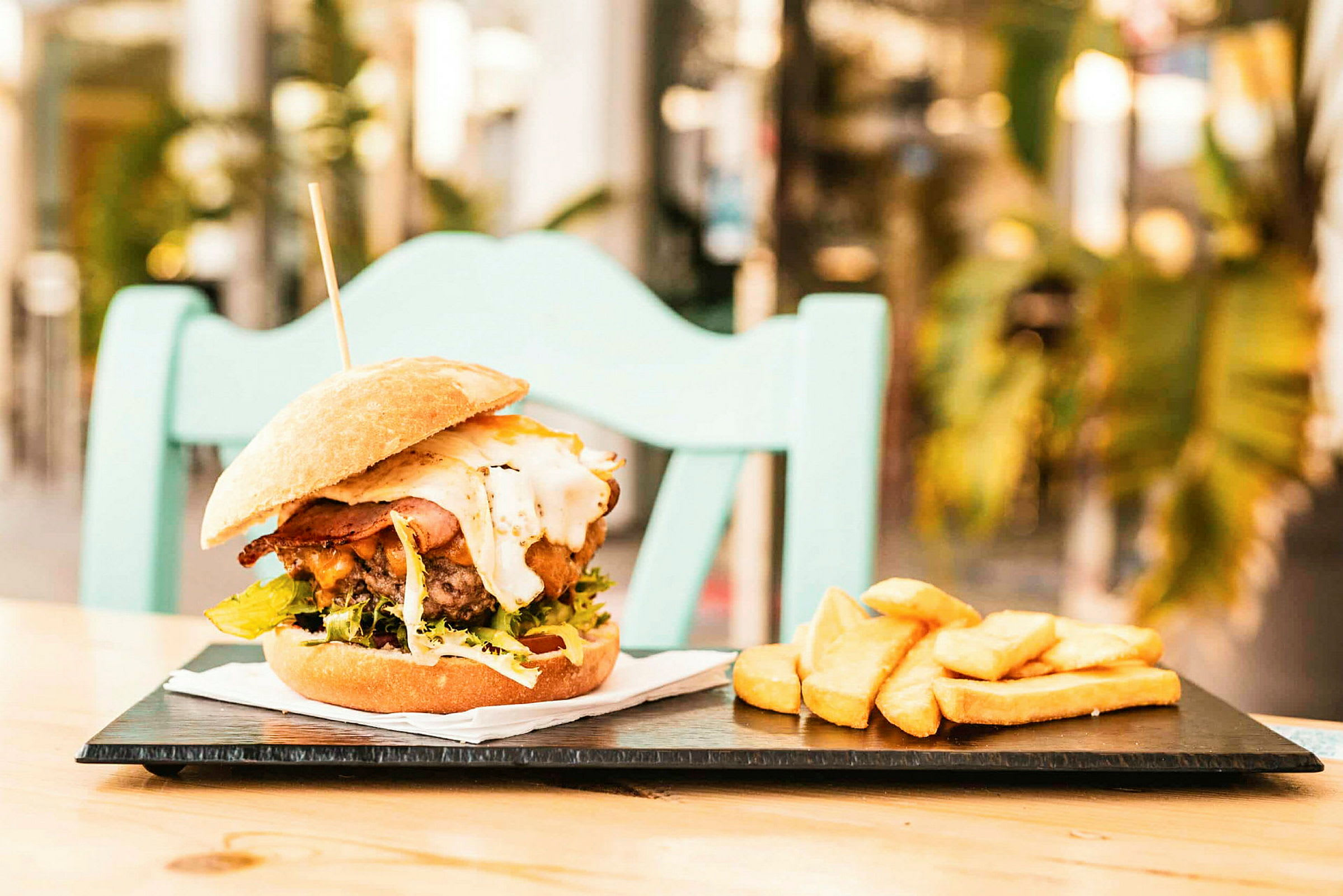 En hamburgare med tillbehör av pommes frites på en tallrik, placerad på ett bord med en stol i bakgrunden. (AI-genererad text)