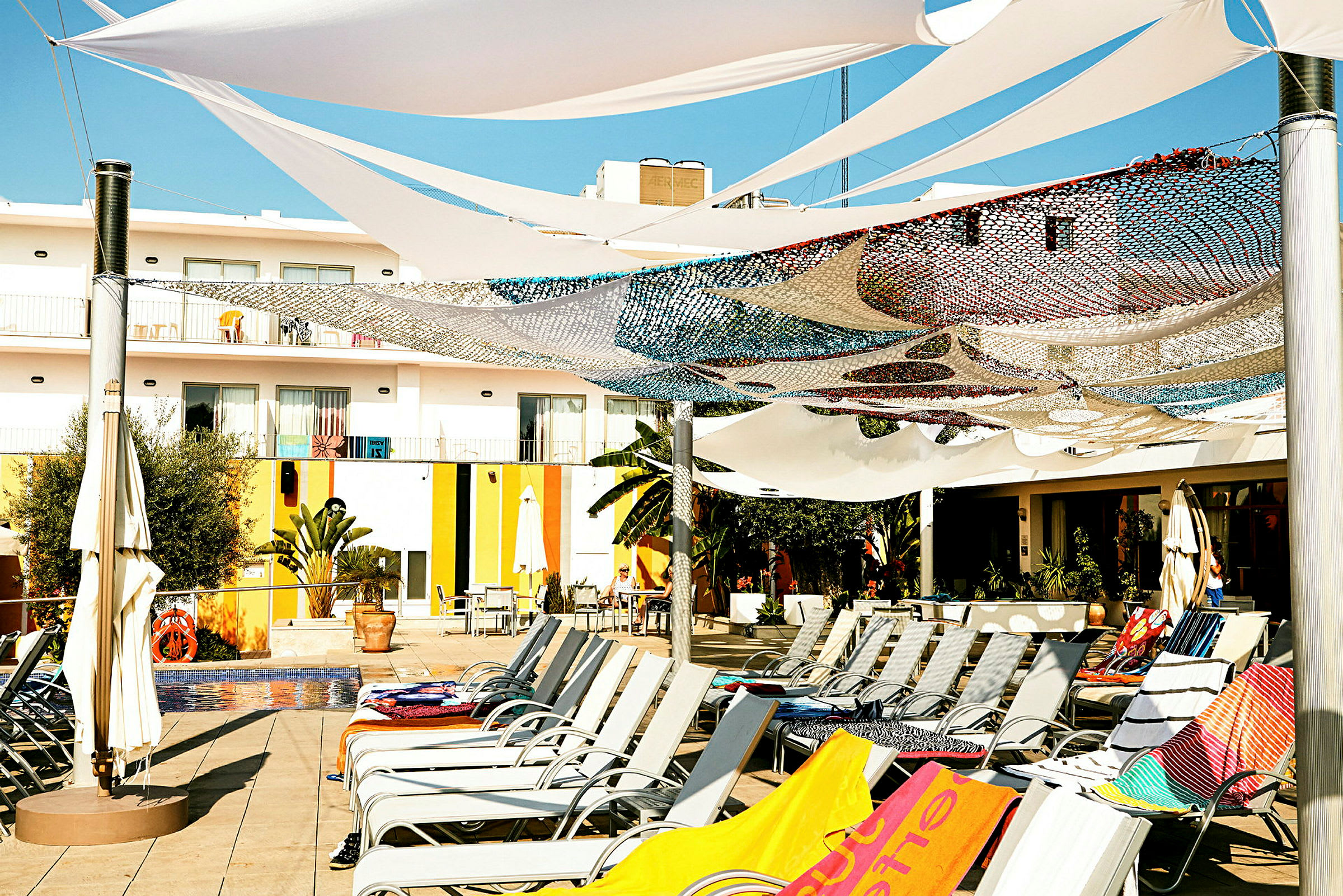 Bild av en solterrass med solstolar och färgglada handdukar under skuggande segel och nät. I bakgrunden syns en modern byggnad med balkonger och växter. (AI-genererad text)