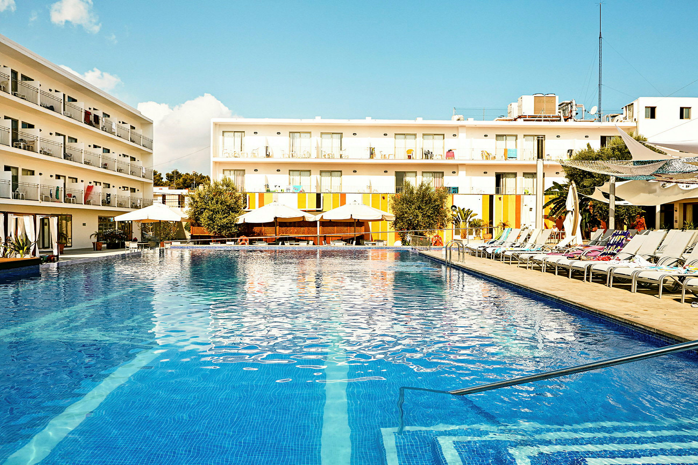 En pool med solstolar och parasoller, framför en modern byggnad med balkonger. (AI-genererad text)