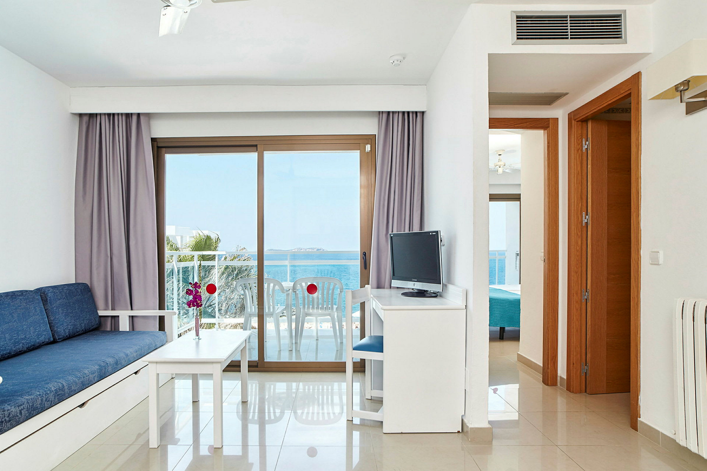 Ett hotellrum med en blå soffa, vitt bord och stol framför ett stort fönster med havsutsikt. En TV står på ett skrivbord. (AI-genererad text)