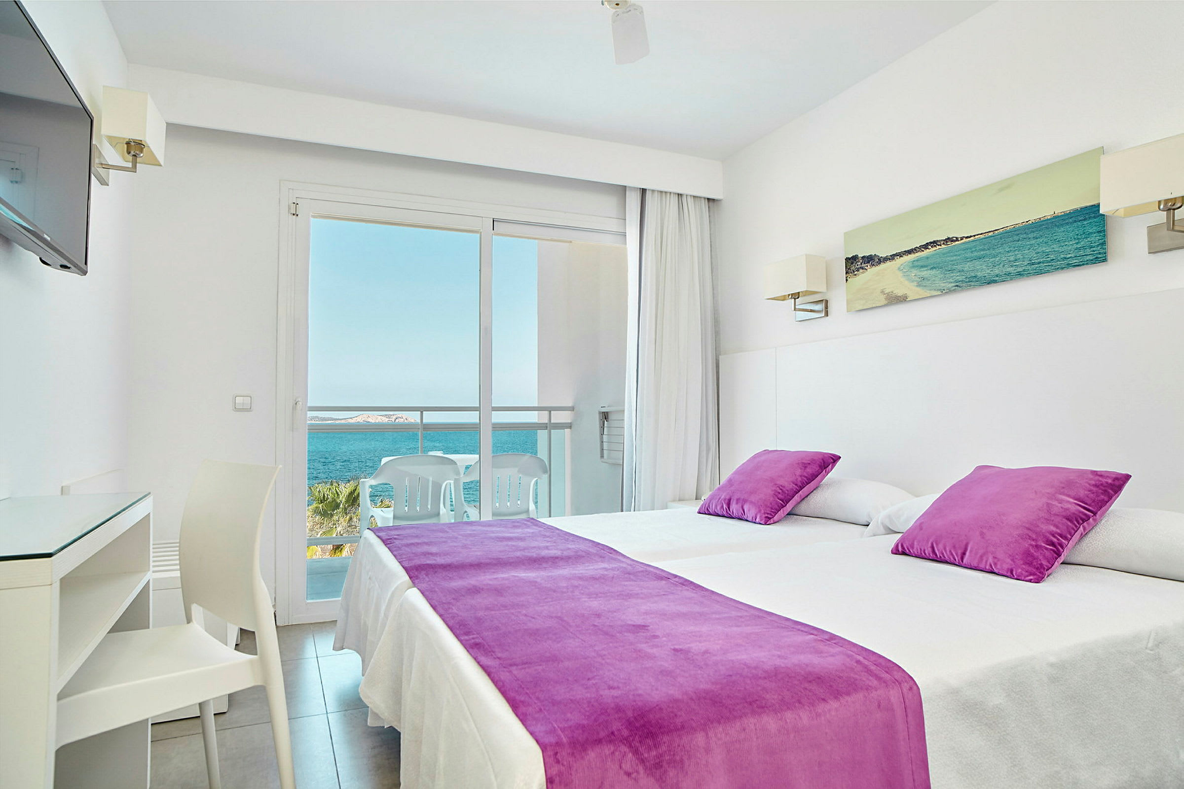 Ett hotellrum med två sängar klädda i vita lakan och lila överkast och kuddar. En balkong med sittmöbler och utsikt mot havet syns genom en glasdörr. (AI-genererad text)