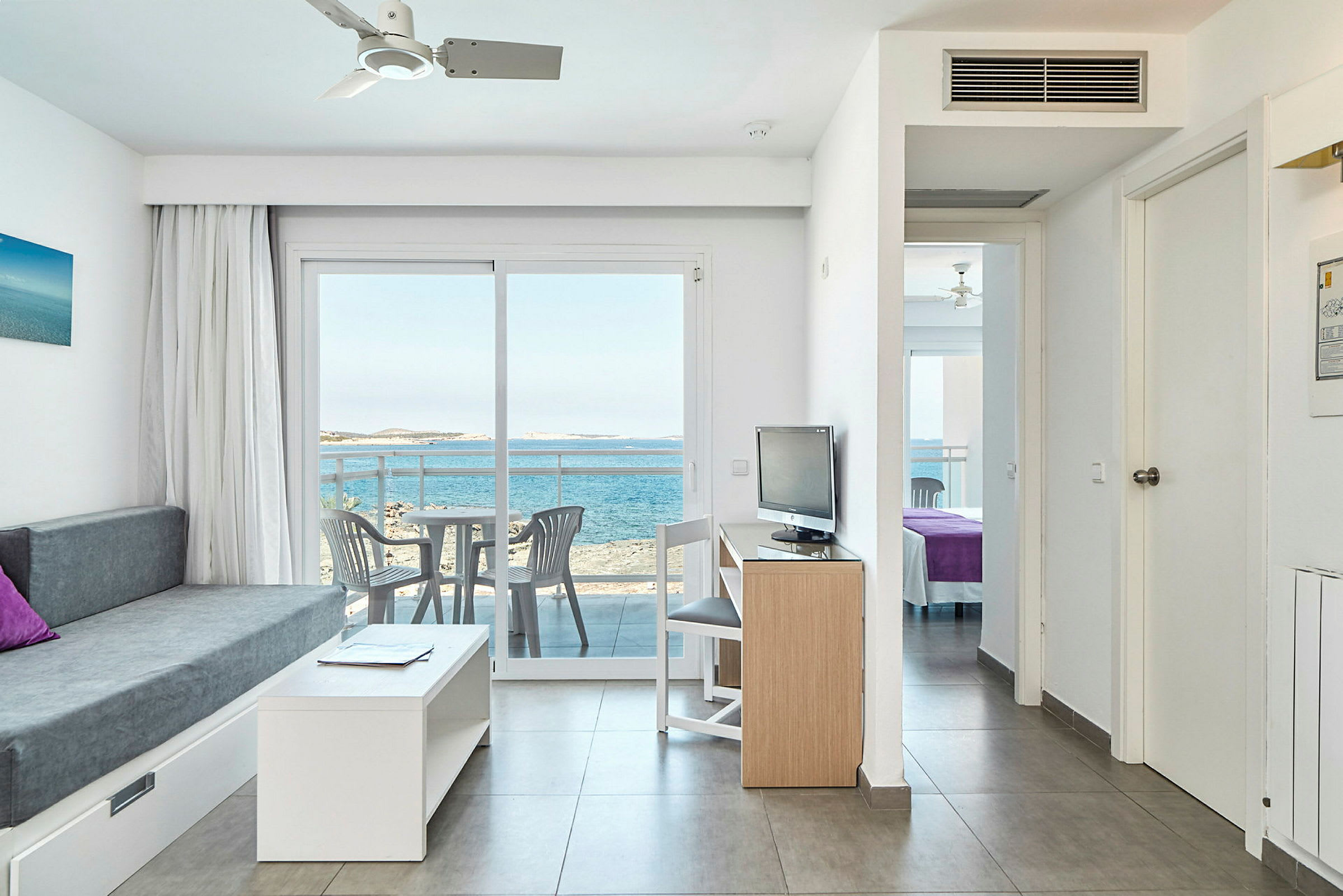 Ett hotellrum med soffa, bord och TV samt en balkong med utsikt över havet. (AI-genererad text)