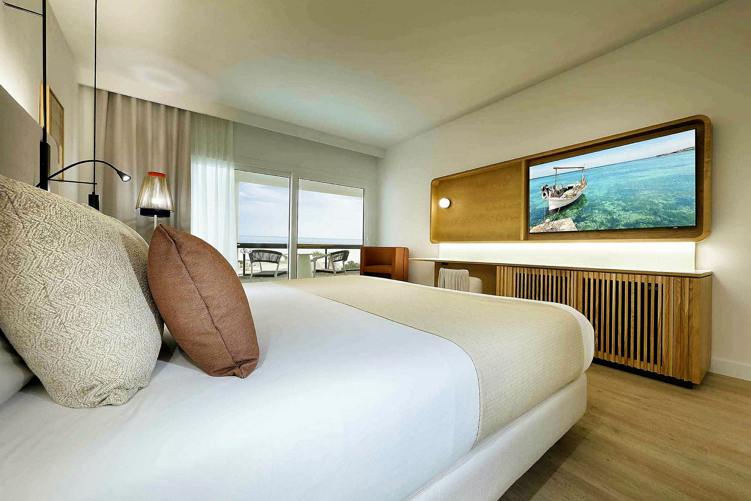Ett hotellrum med en stor säng, dekorativa kuddar, ett skrivbord och ett stort fönster som leder till en balkong. (AI-genererad text)