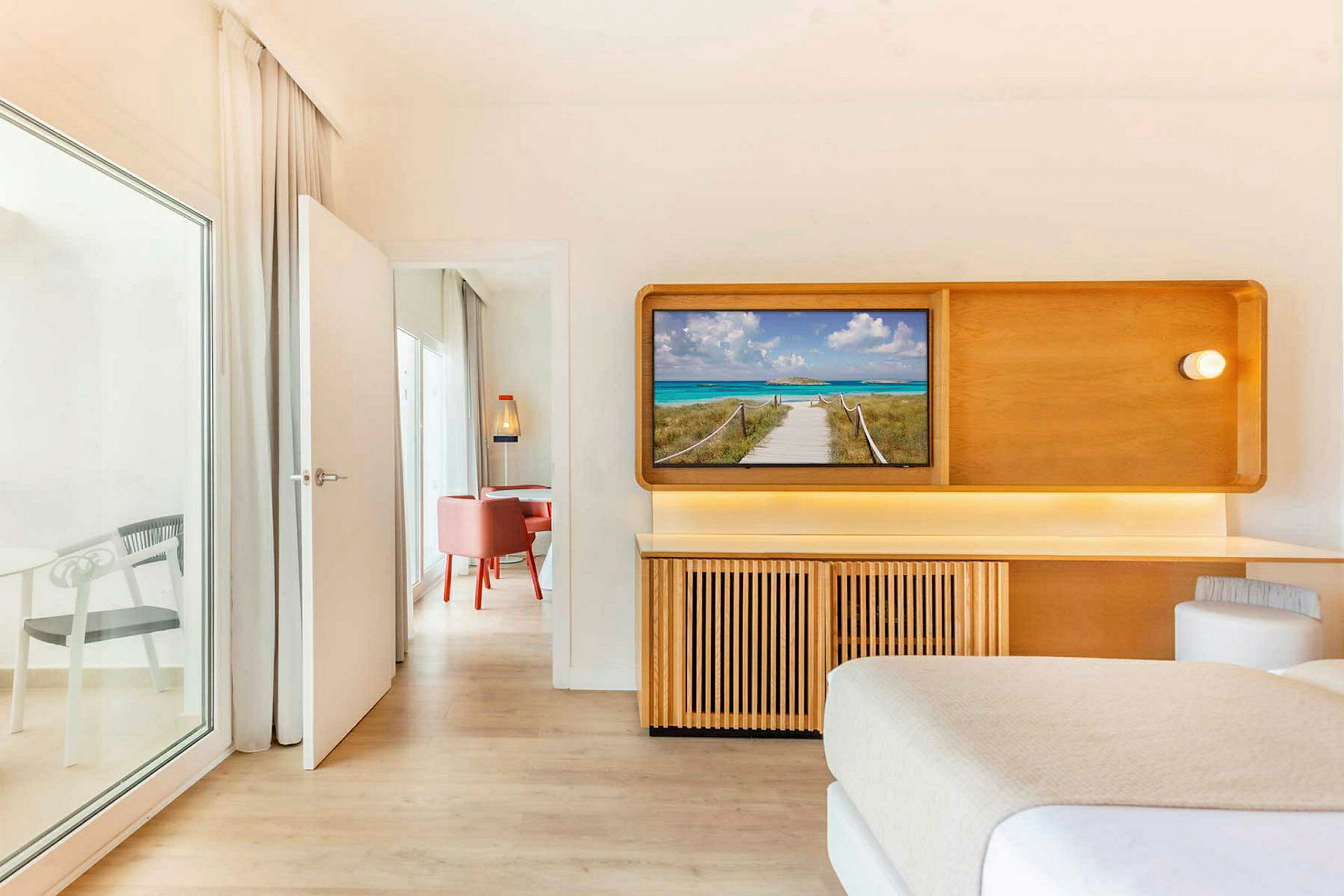 Ett hotellrum med en TV som visar en strandpromenad. En dörr leder till ett angränsande rum med röda stolar. (AI-genererad text)