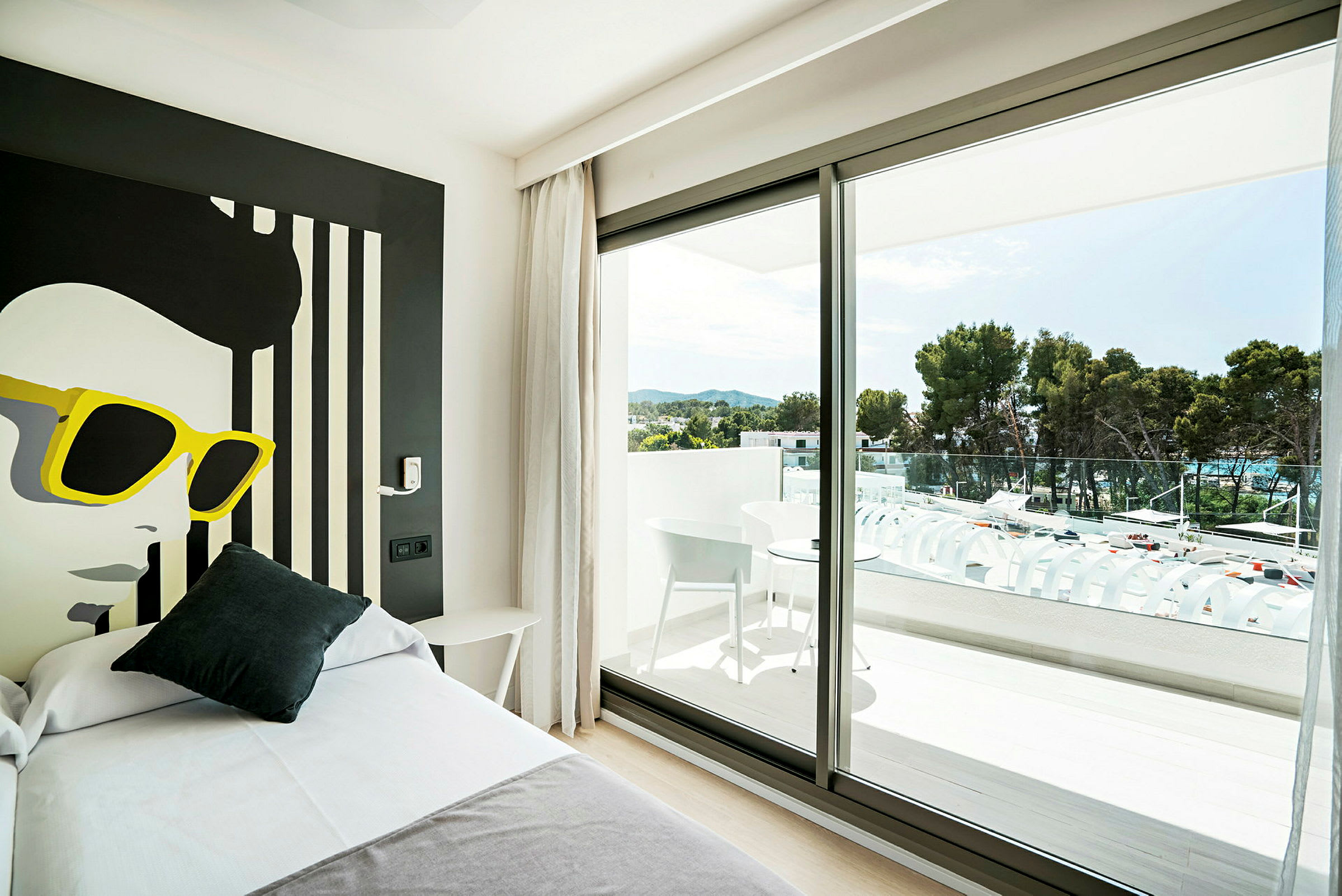 Ett hotellrum med en modern inredning, en säng i förgrunden och en stor fönsterruta som leder ut till en balkong med möbler och utsikt över grönskande träd. (AI-genererad text)