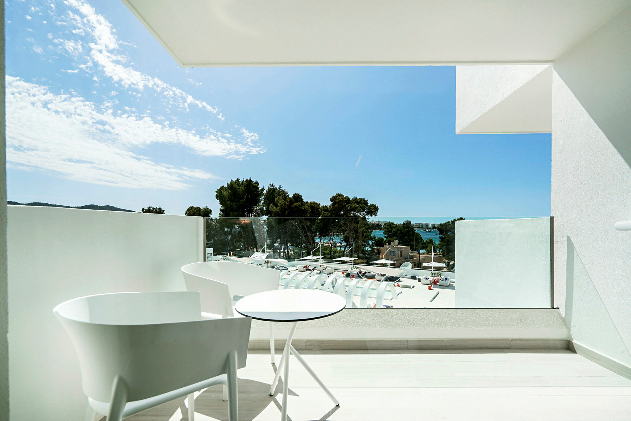 En balkong med vita stolar och ett runt bord som vetter mot en utsikt med träd, byggnader och hav. (AI-genererad text)
