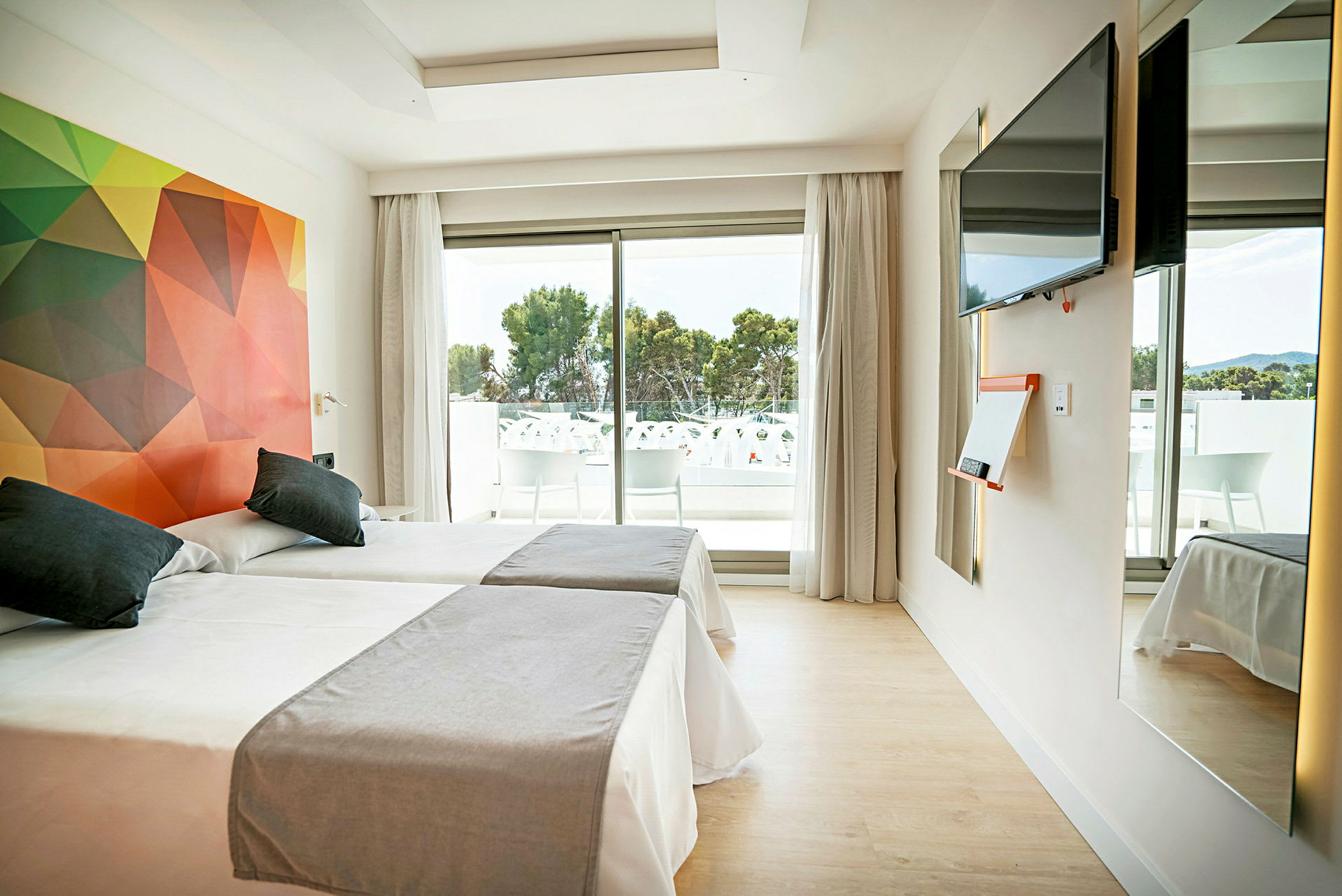 Ett hotellrum med två enkelsängar, färgglada väggdekorationer, en TV på väggen, ett stort fönster som öppnar mot en balkong med sittplatser och utsikt över träd. (AI-genererad text)