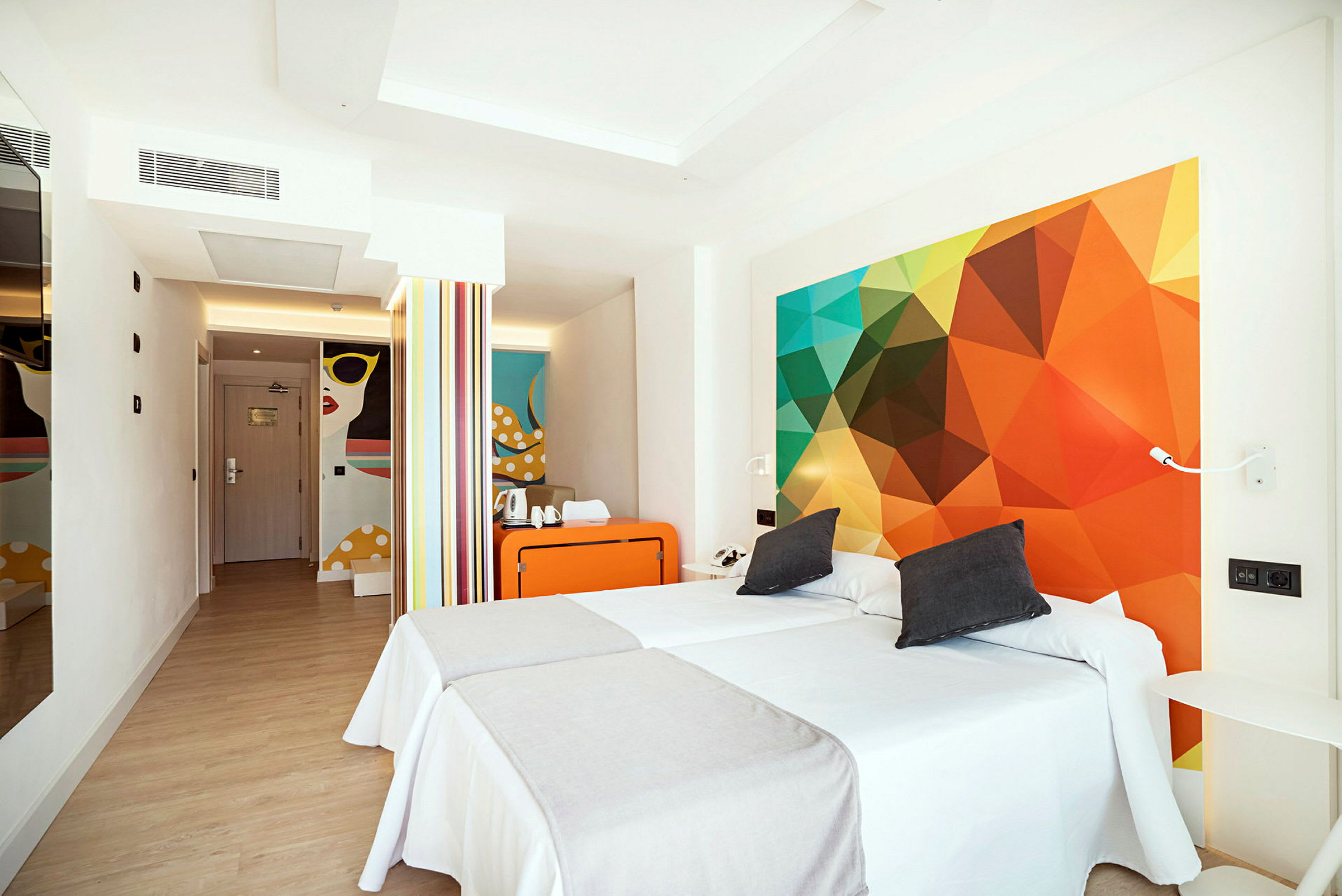 Ett modernt hotellrum med två enkelsängar, färgglada väggdekorationer och en liten sittplats. (AI-genererad text)