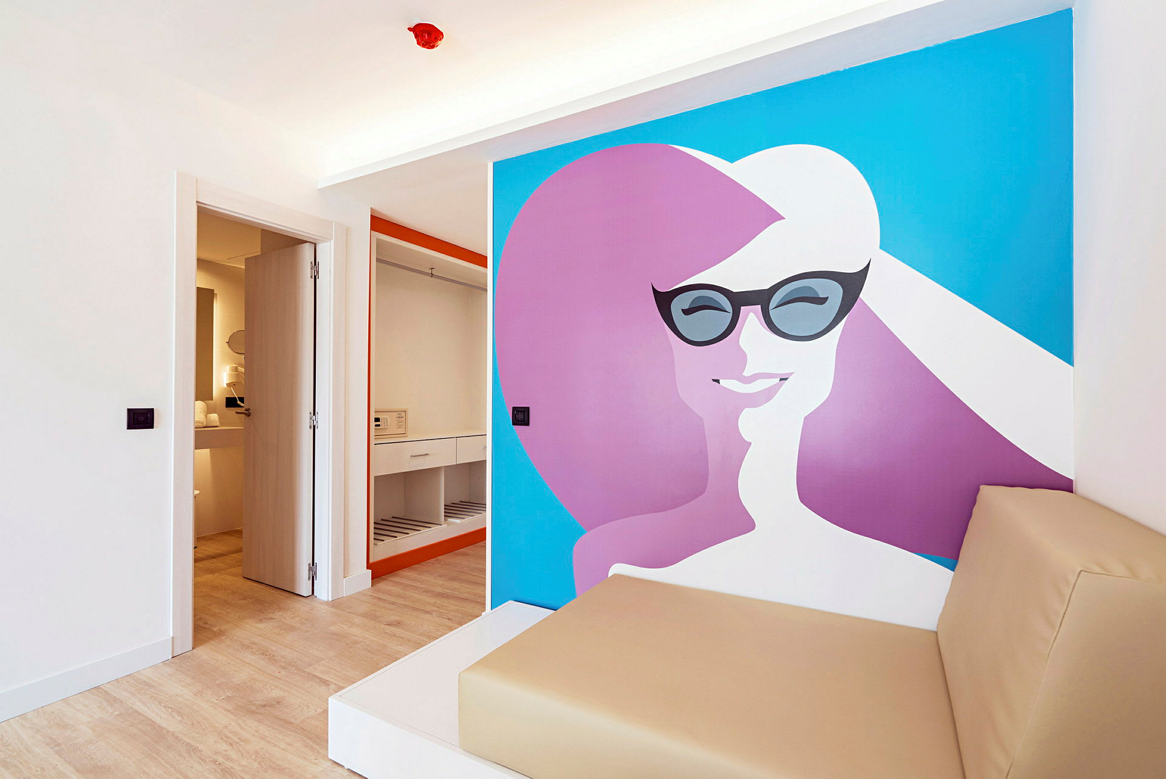 Bild av ett hotellrum med öppen garderob, badrumsdörr och dekorativ väggmålning med färgglada former. En beige sittmöbel syns i förgrunden. (AI-genererad text)