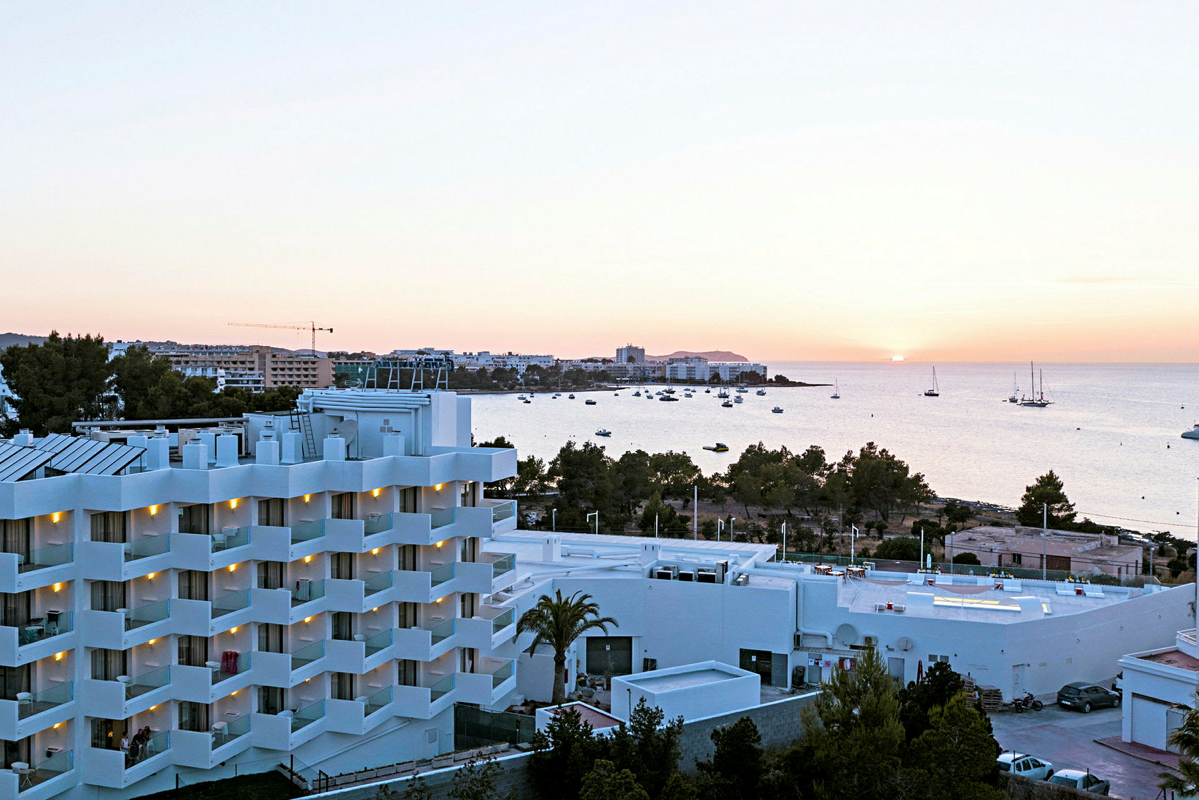 En vy över ett hotell med vita fasader och balkonger, omgivna av grönska, med havet och en solnedgång i bakgrunden. (AI-genererad text)