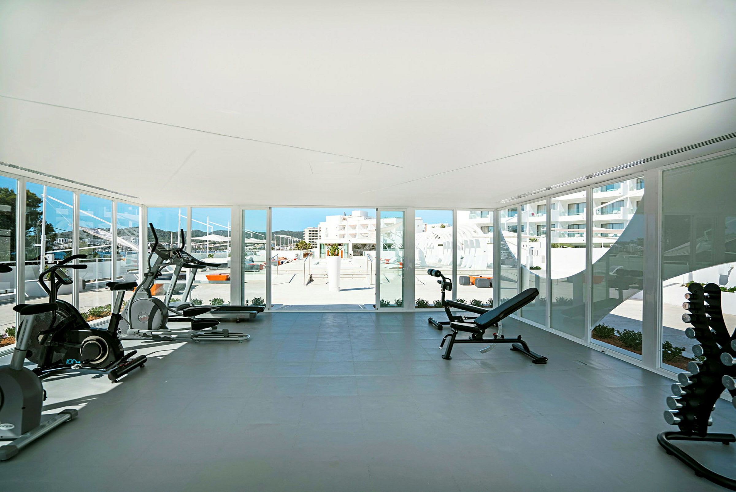 Gym med träningsutrustning och utsikt genom stora fönster mot en solig utomhusmiljö. (AI-genererad text)
