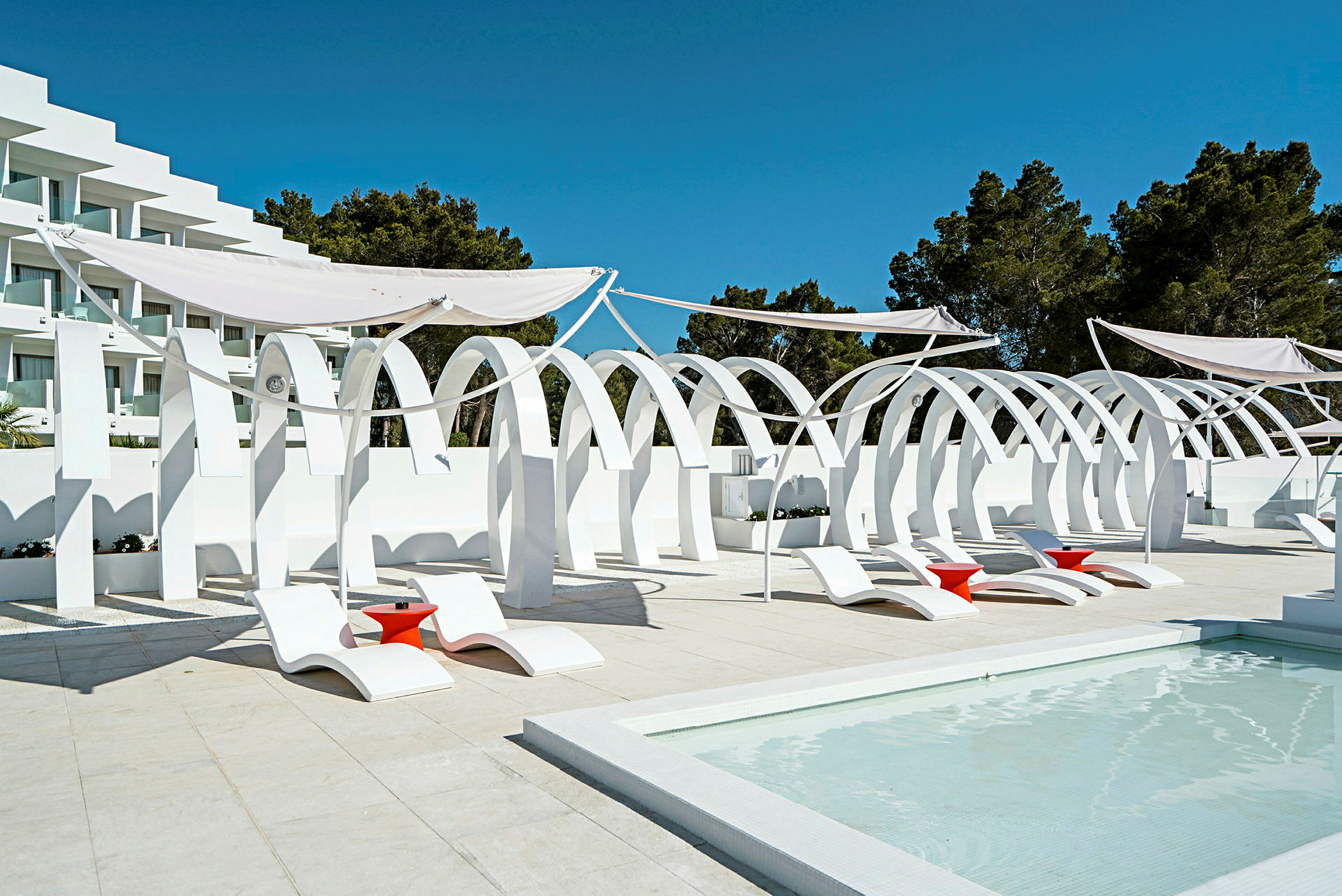 Vit arkitektur med bågar, samt solstolar och röda bord vid en pool. (AI-genererad text)