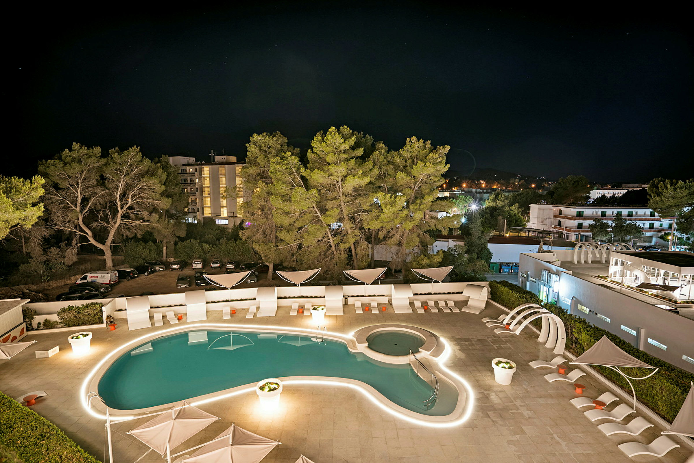 En nattlig vy över en hotellpool omgiven av solstolar och belysning, med träd och byggnader i bakgrunden. (AI-genererad text)