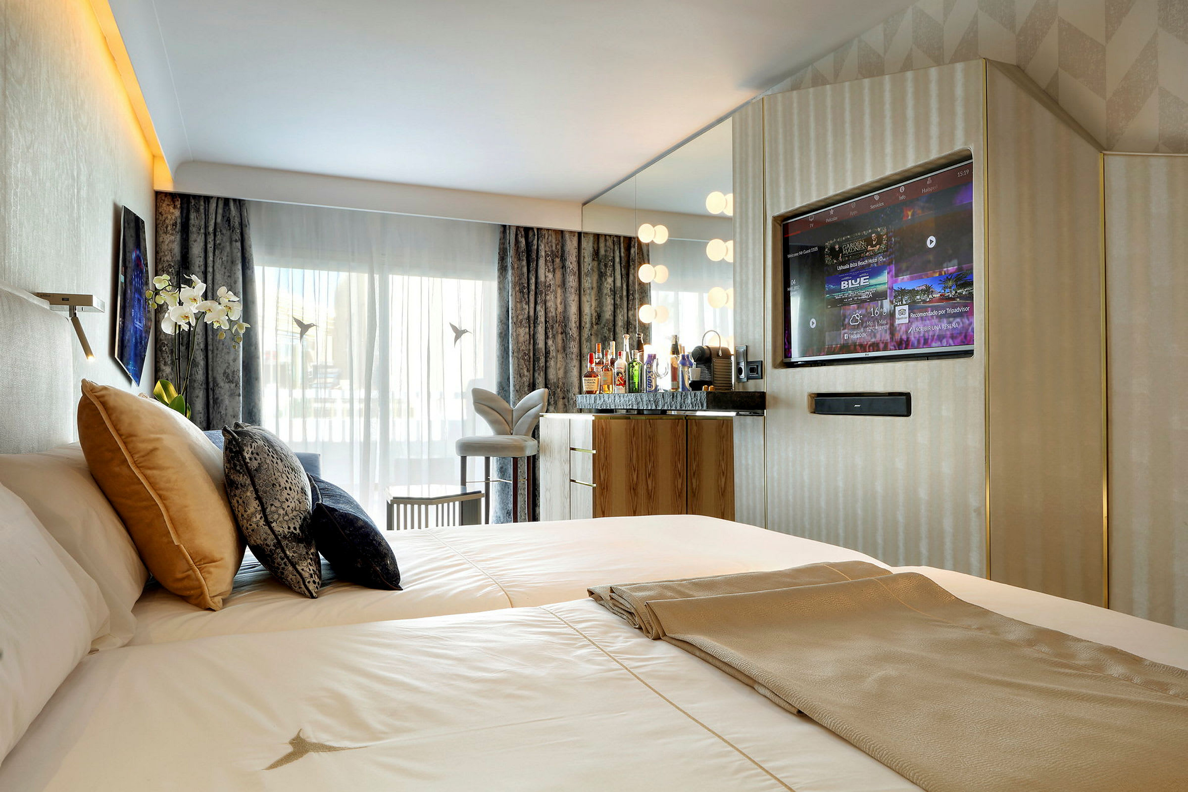 Ett hotellrum med en bred säng, flera kuddar, en TV inbyggd i väggen och en speglad bänk med drycker och lampor. Stora fönster med gardiner och en dekorativ stol är också synliga. (AI-genererad text)
