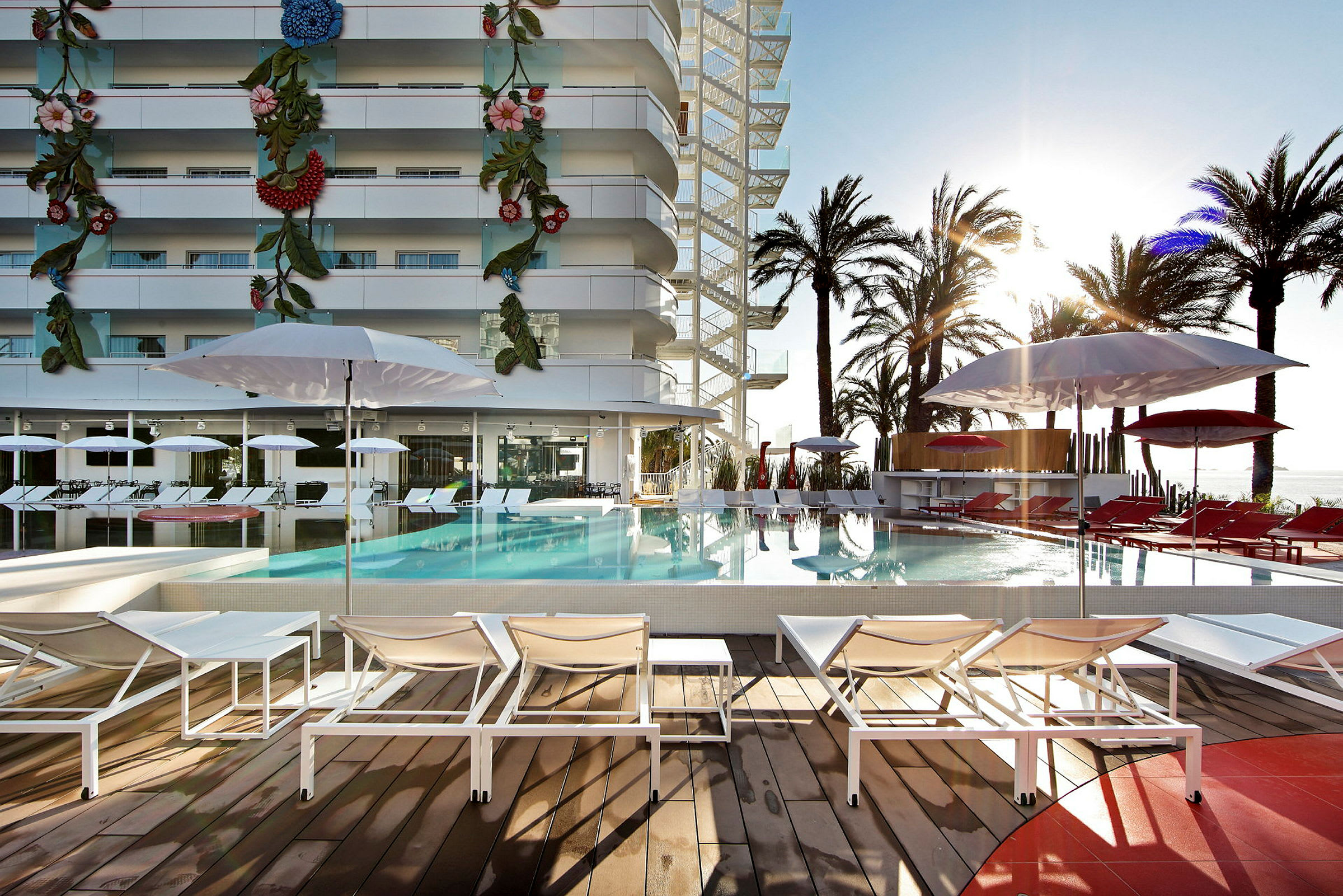 Utomhuspoolområde med solstolar och parasoller kring en pool och en modern byggnad i bakgrunden. (AI-genererad text)