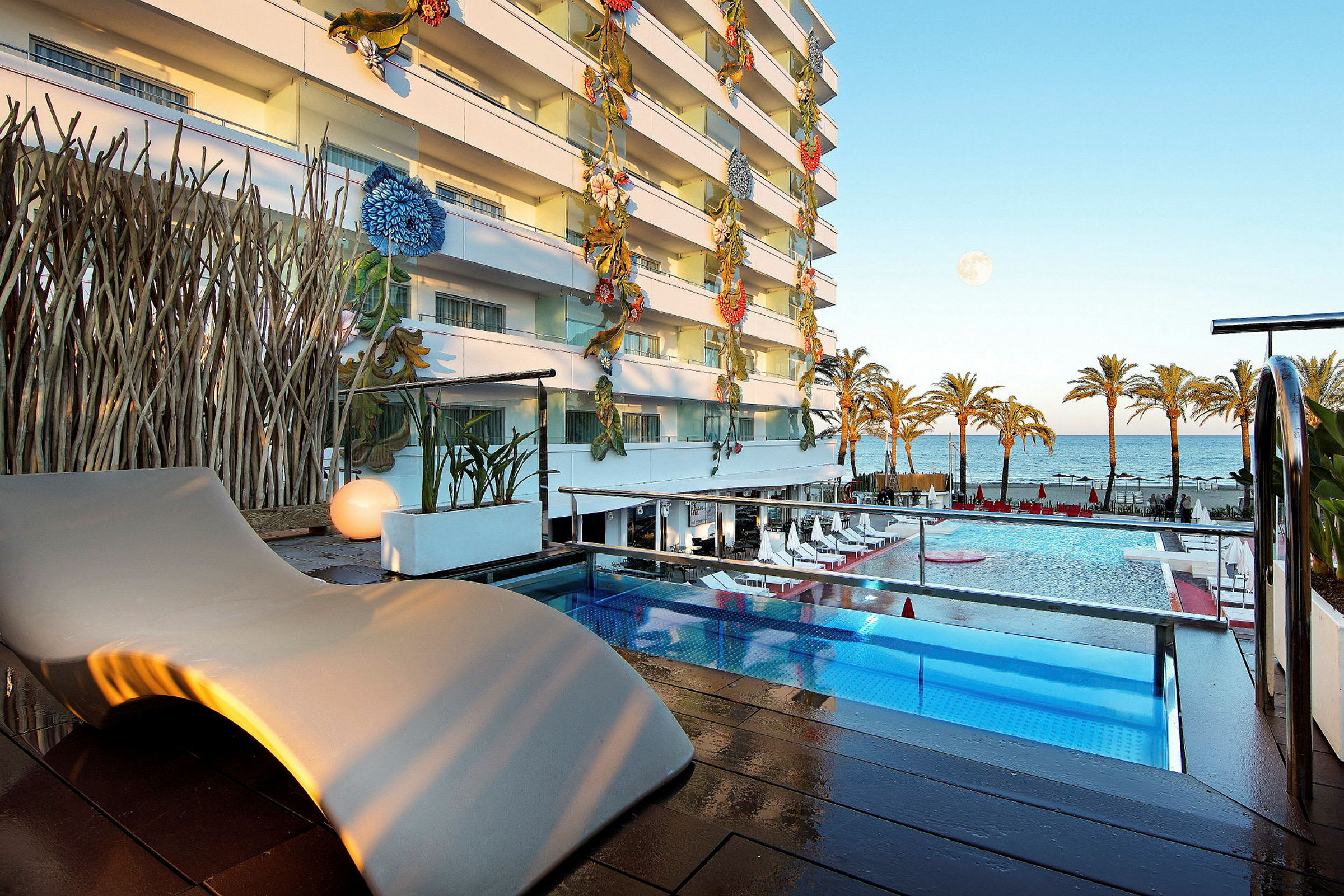 Ett hotellområde med en pool, soldäck, och utsikt över havet. Byggnaden har färgglada blomdekorationer längs fasaden. (AI-genererad text)