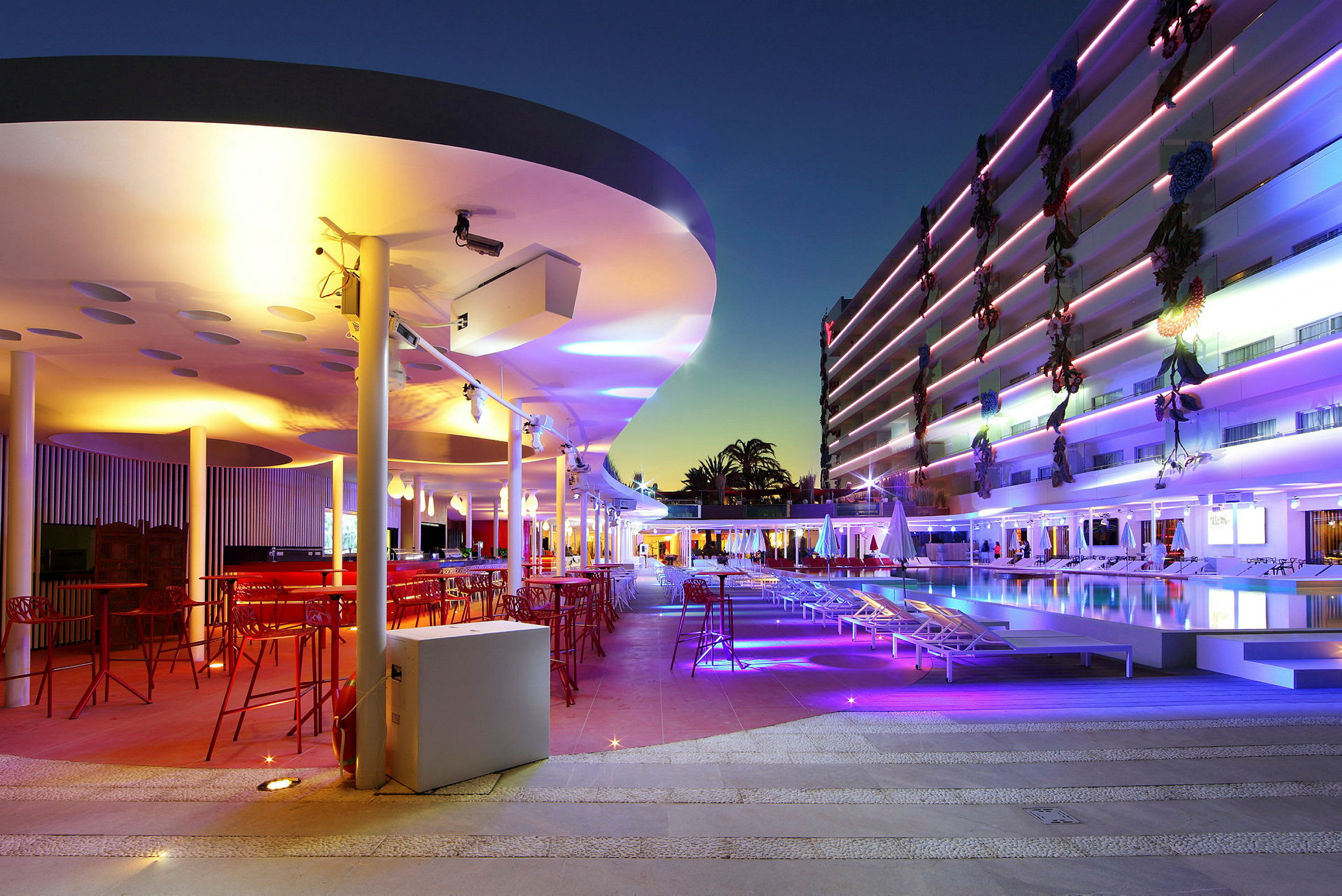 Ett modernt utomhusområde med bar och pool, upplyst av färgglada lampor under kvällstid. (AI-genererad text)