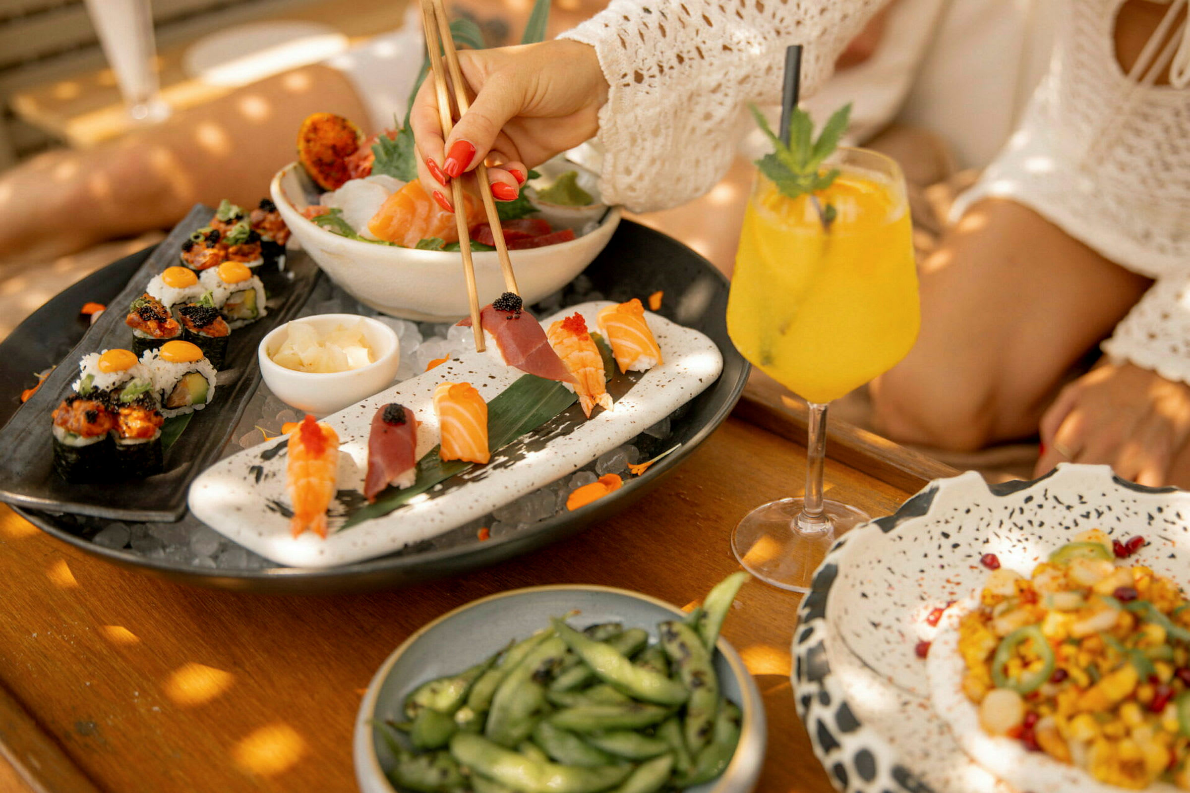 En tallrik med sushi och sashimi tillsammans med en gul dryck och en skål sojabönor stående på ett träbord. (AI-genererad text)