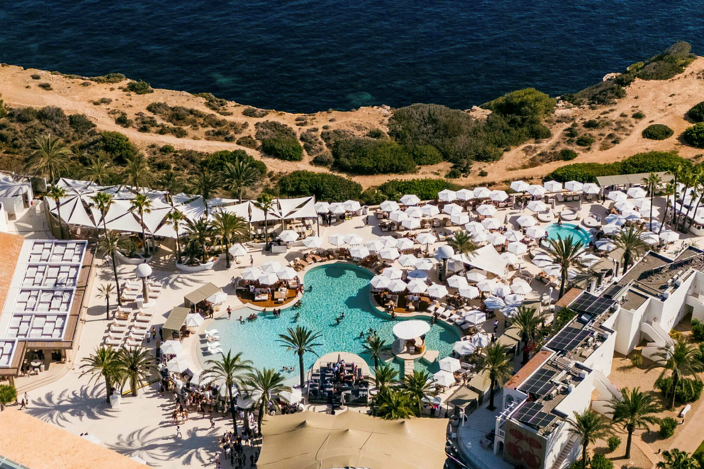 Flygfoto av en semesterresort med flera pooler och vita parasoller, angränsande till klippor och hav. (AI-genererad text)