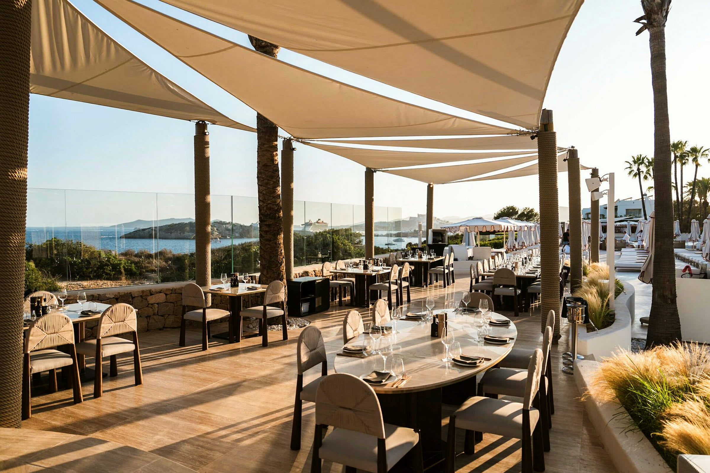 Restaurang med bord och stolar på en terrass nära havet, skuggad av segelduk. (AI-genererad text)