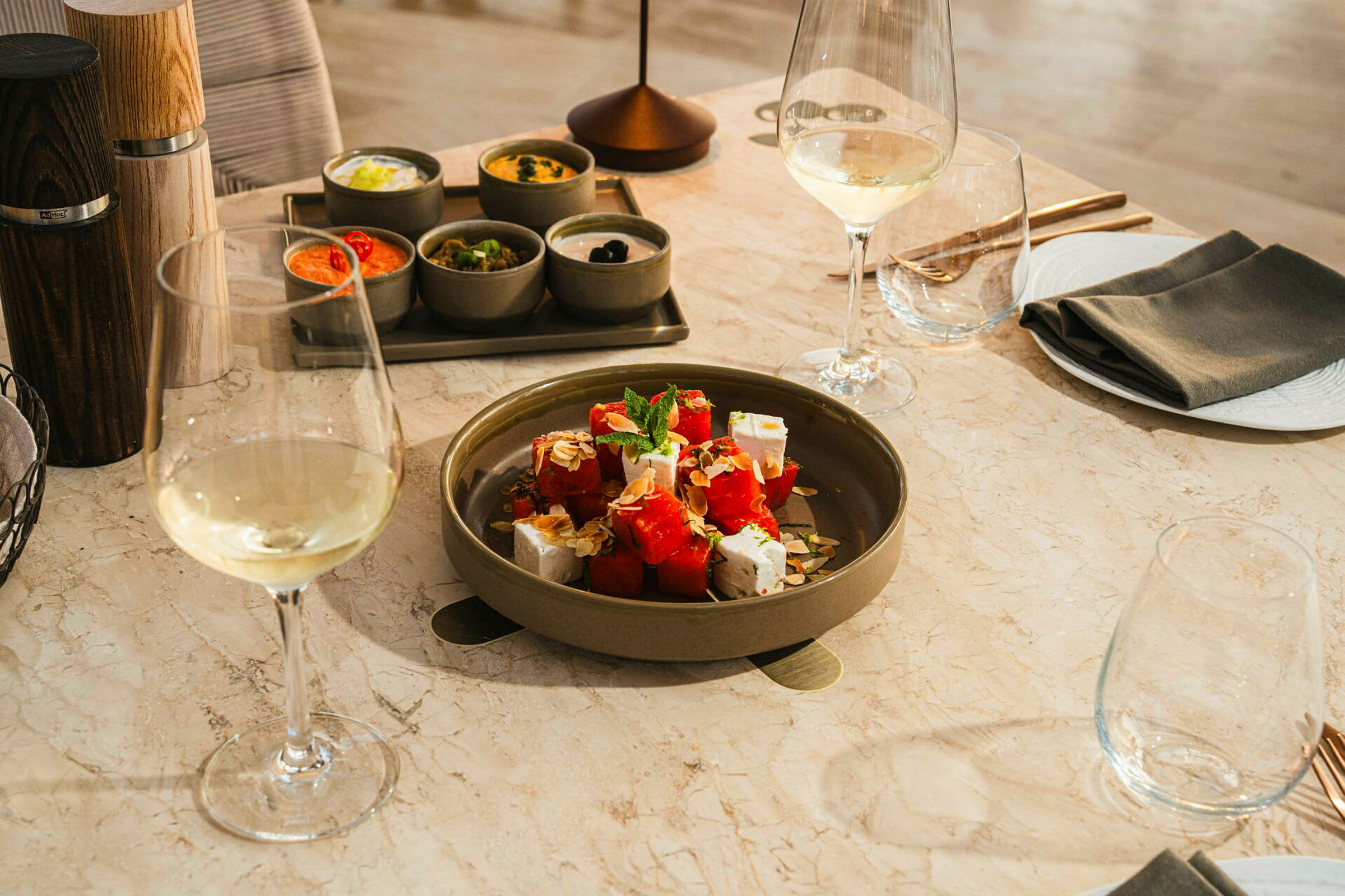 Ett bord med en tallrik innehållande sallad med vattenmelon och fetaost, omgiven av olika tillbehör i små skålar samt glas med vitt vin. (AI-genererad text)