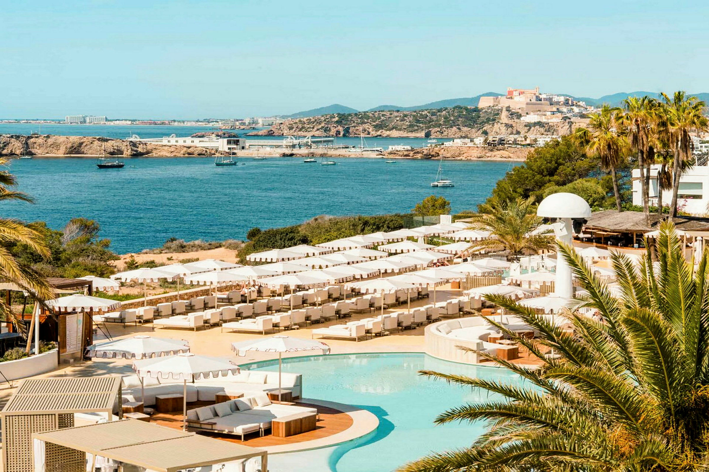 Bild av en kustlinje med ett hotellkomplex innehållande en pool och solstolar under parasoller. I bakgrunden ses havet med båtar och en stad på en kulle. (AI-genererad text)