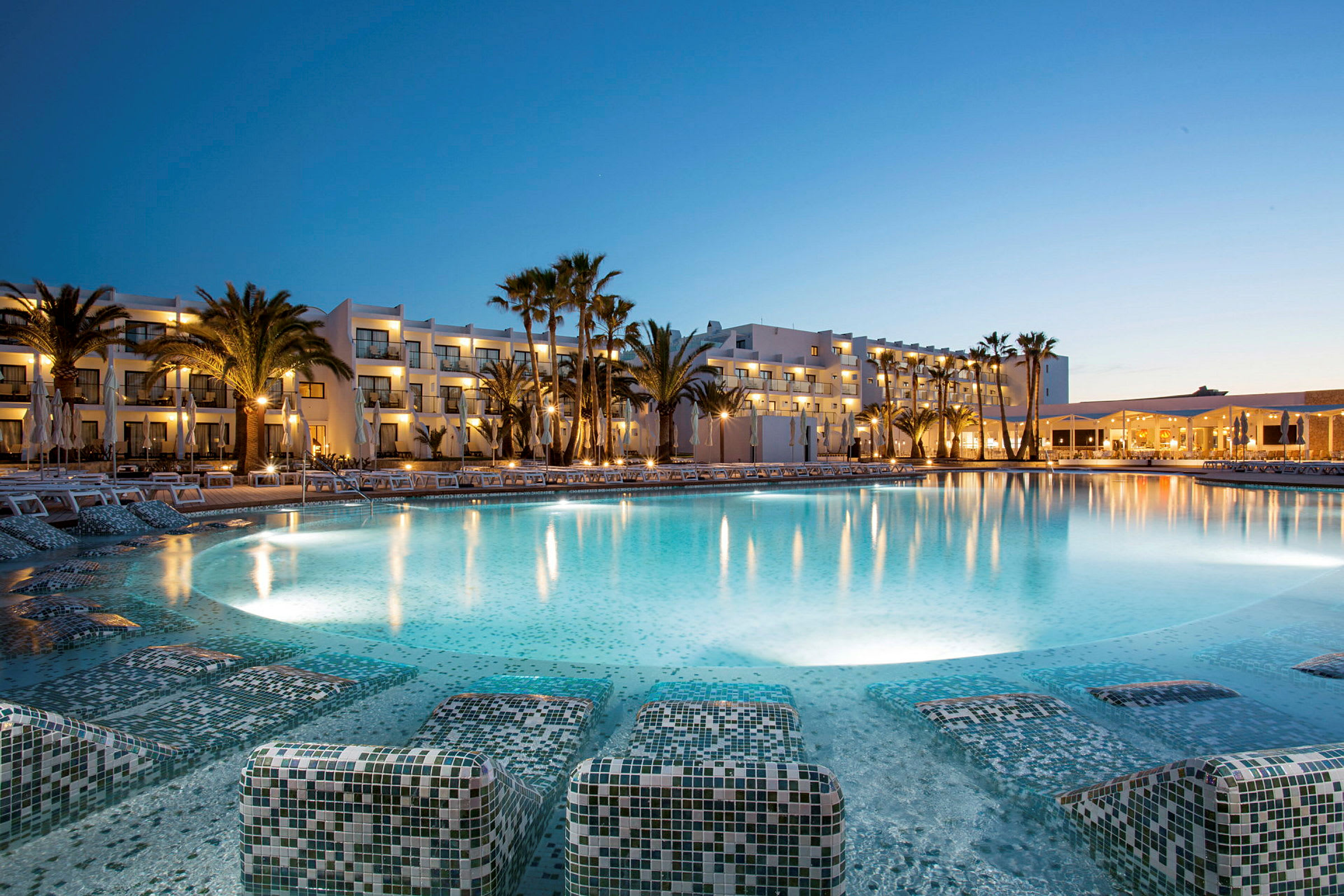 Bild av en stor pool framför ett hotellkomplex med upplysta fönster vid skymning. (AI-genererad text)