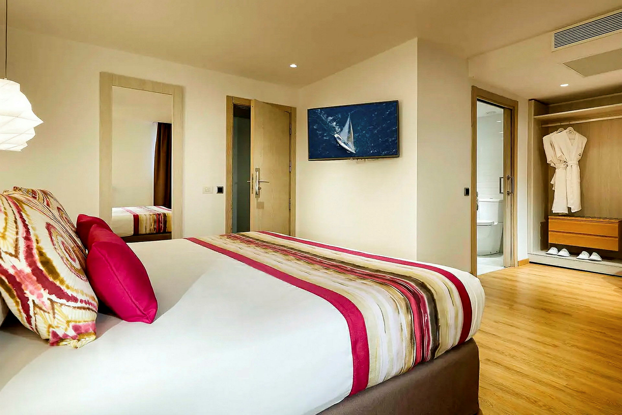Ett hotellrum med modern inredning, en säng med färgglada kuddar och ett täcke, en väggmonterad TV, ett badrum i anslutning och en garderob med badrockar. (AI-genererad text)