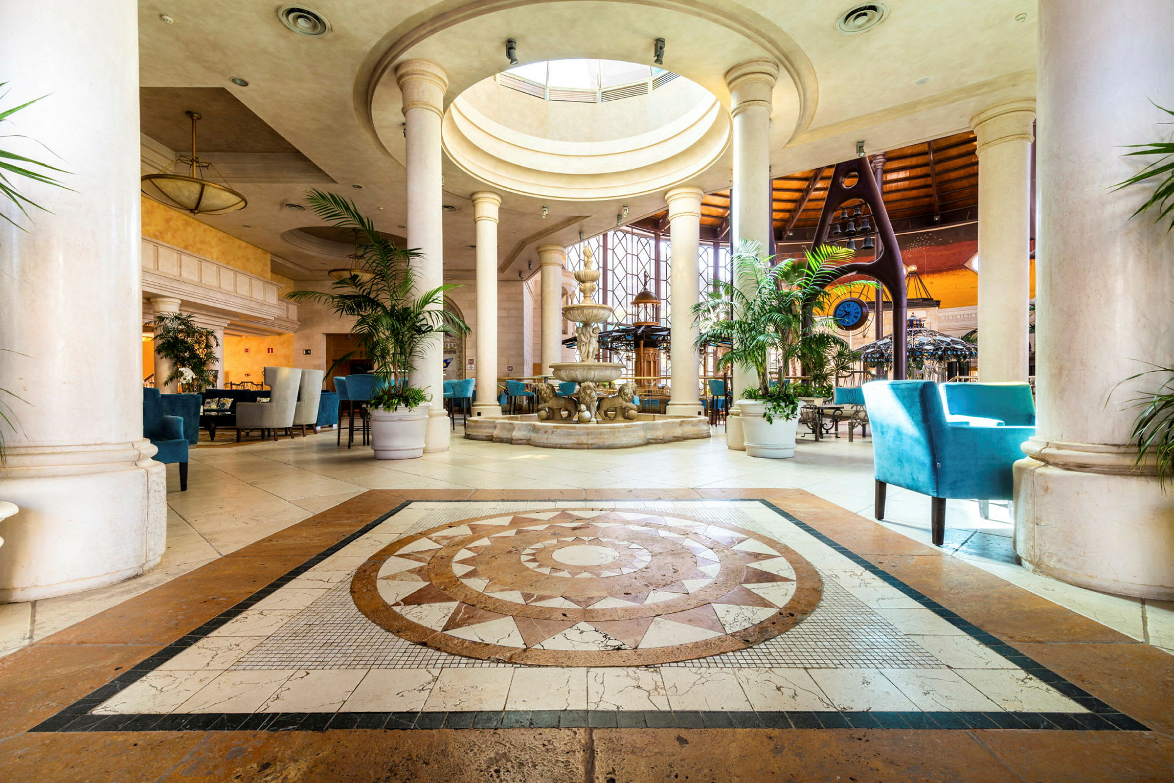Interiör av en hotellobby med marmorgolv, pelare, en fontän och blå stolar. (AI-genererad text)