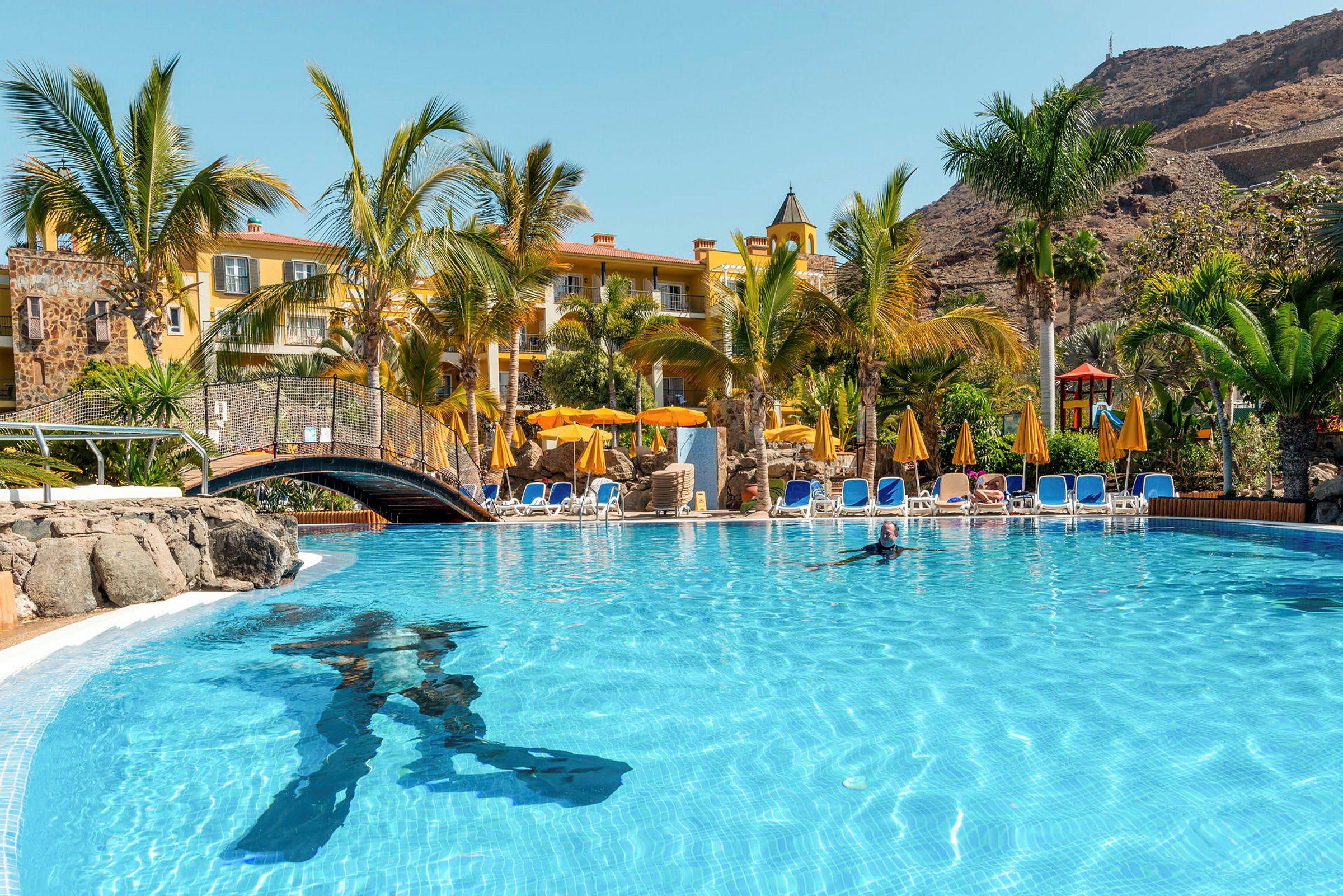 En pool med solstolar och parasoller samt en byggnad i bakgrunden. (AI-genererad text)