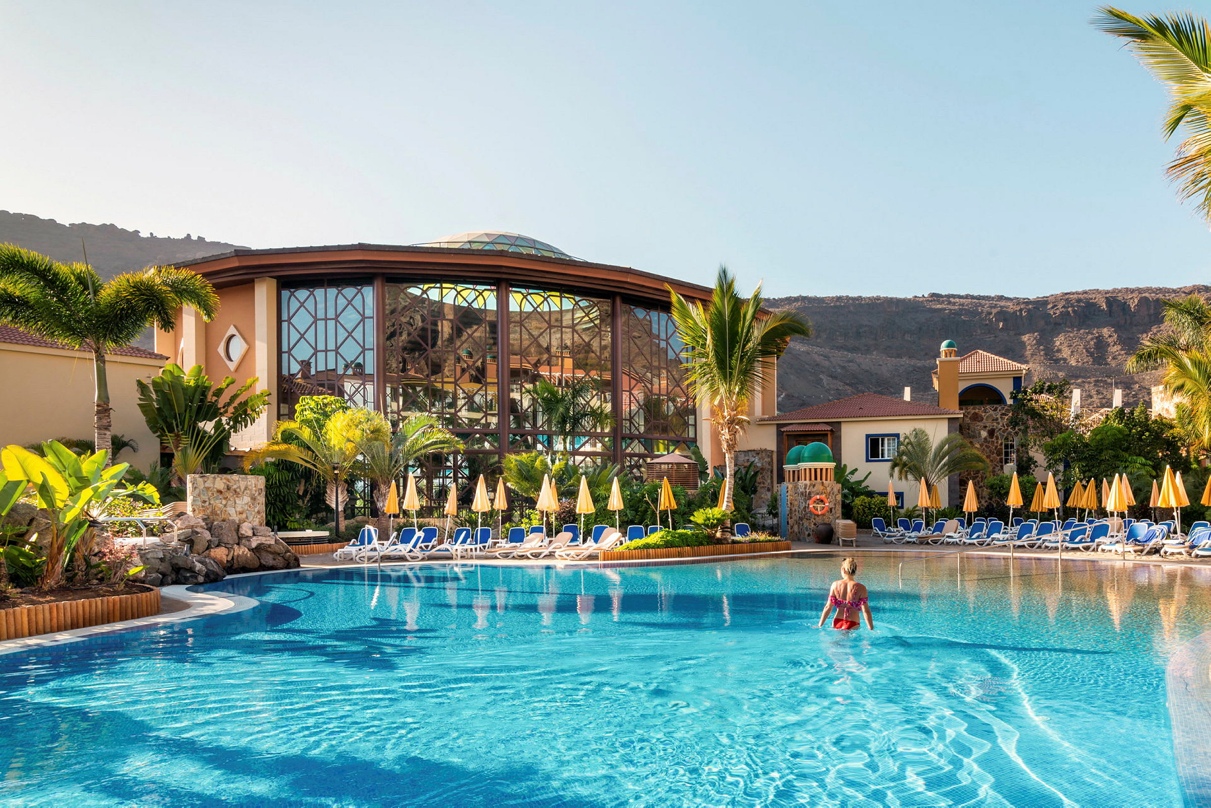 En kvinna står i en pool vid ett hotell med solstolar och parasoller runtom. (AI-genererad text)