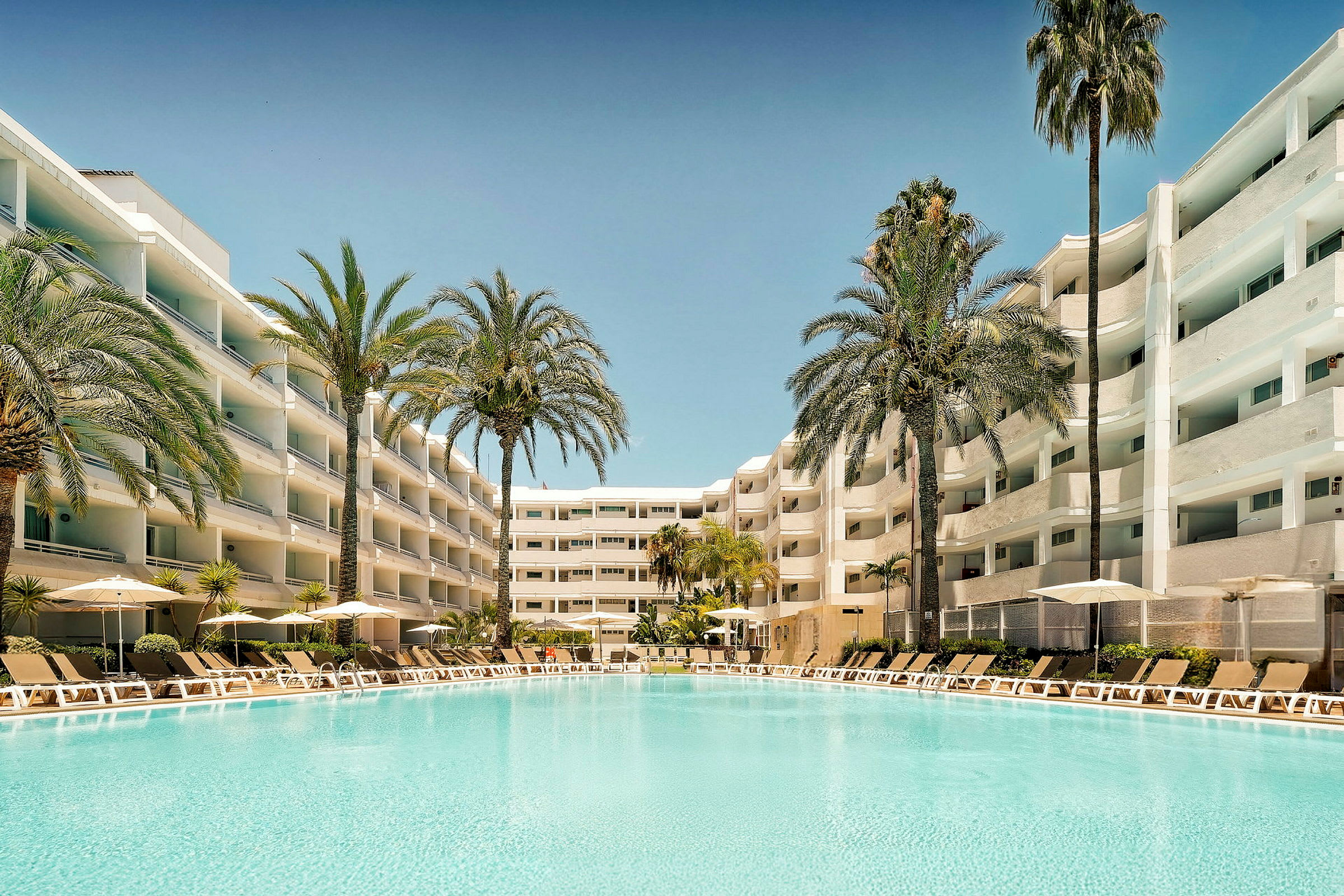 En stor pool med klart vatten omgiven av solstolar och parasoller, med vita hotellbyggnader i bakgrunden och palmer runt omkring. (AI-genererad text)