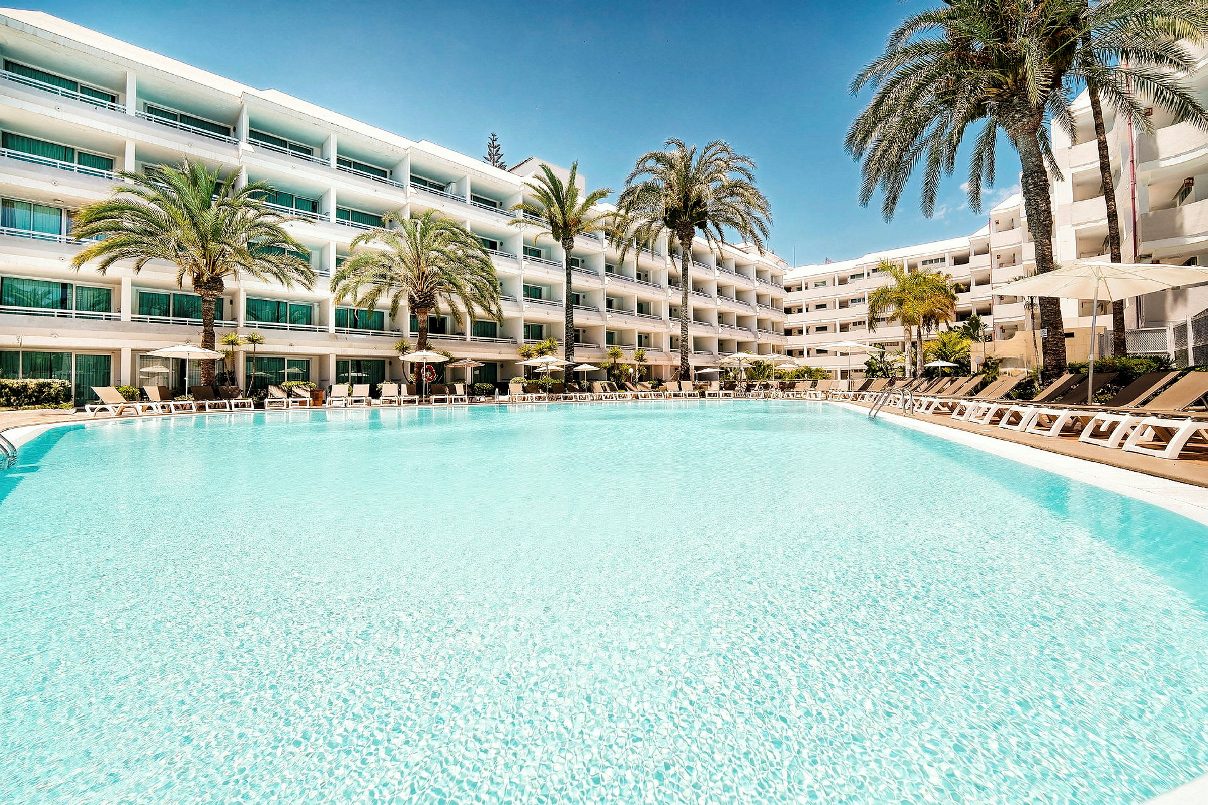 Ett hotellkomplex med flera våningar syns i bakgrunden, och framför byggnaden finns en stor, ljusblå pool omgiven av solstolar och parasoller. (AI-genererad text)
