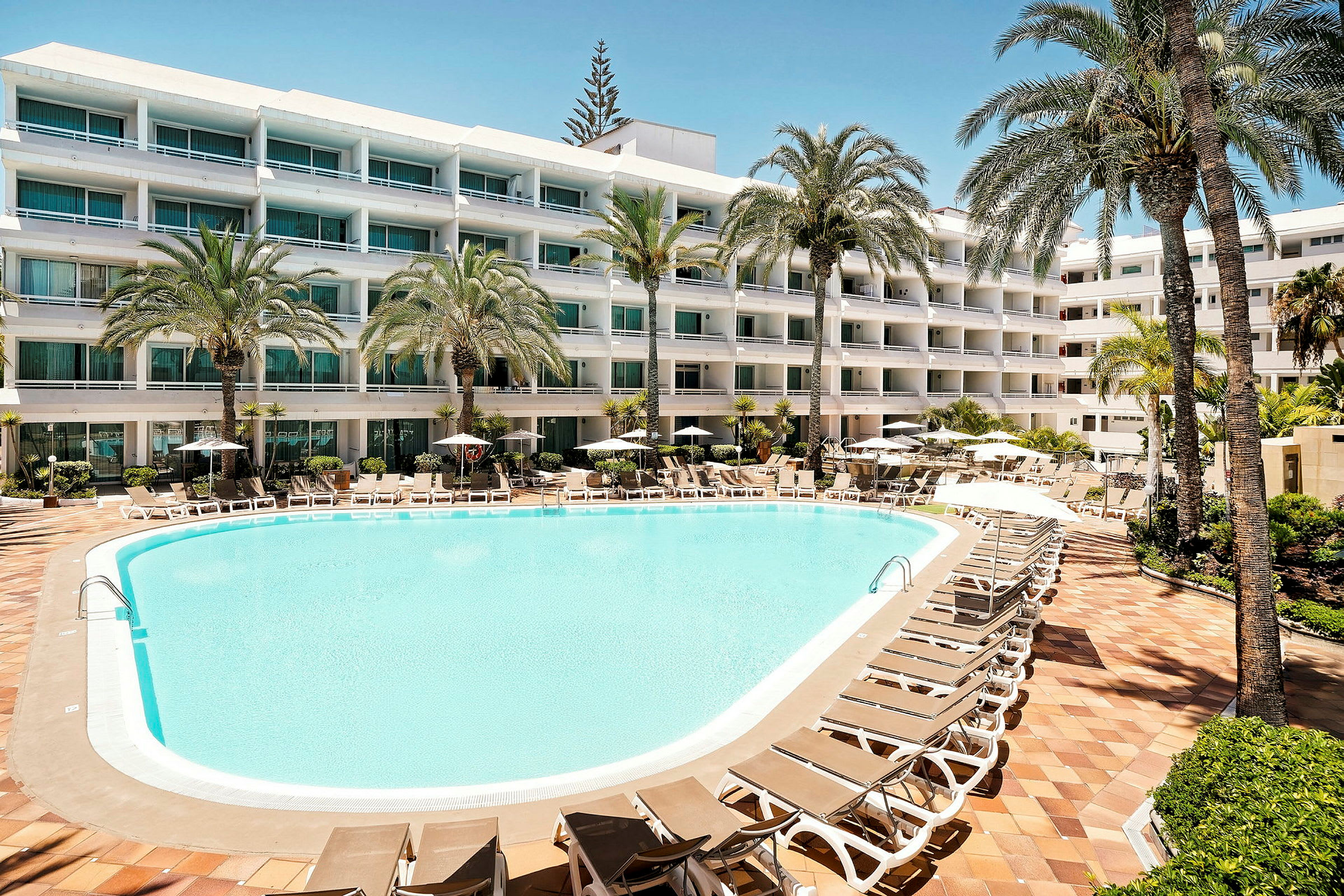 En hotellpool med solstolar och parasoller omgiven av lummiga palmer, samt en flerfaldig byggnad med balkonger i bakgrunden. (AI-genererad text)