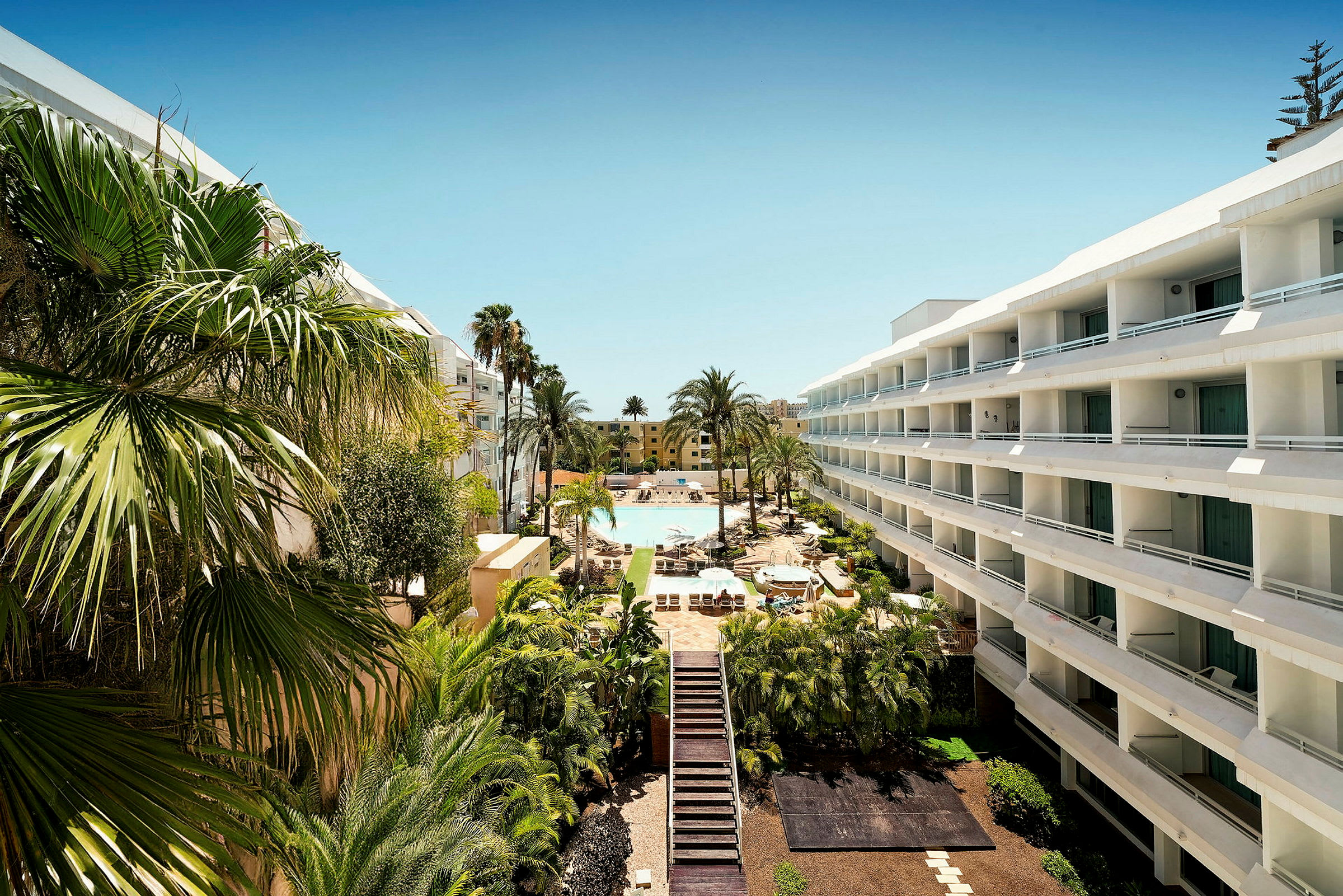 En hotellanläggning med byggnader på båda sidor och en pool i mitten, omgiven av palmer och frodig grönska. (AI-genererad text)