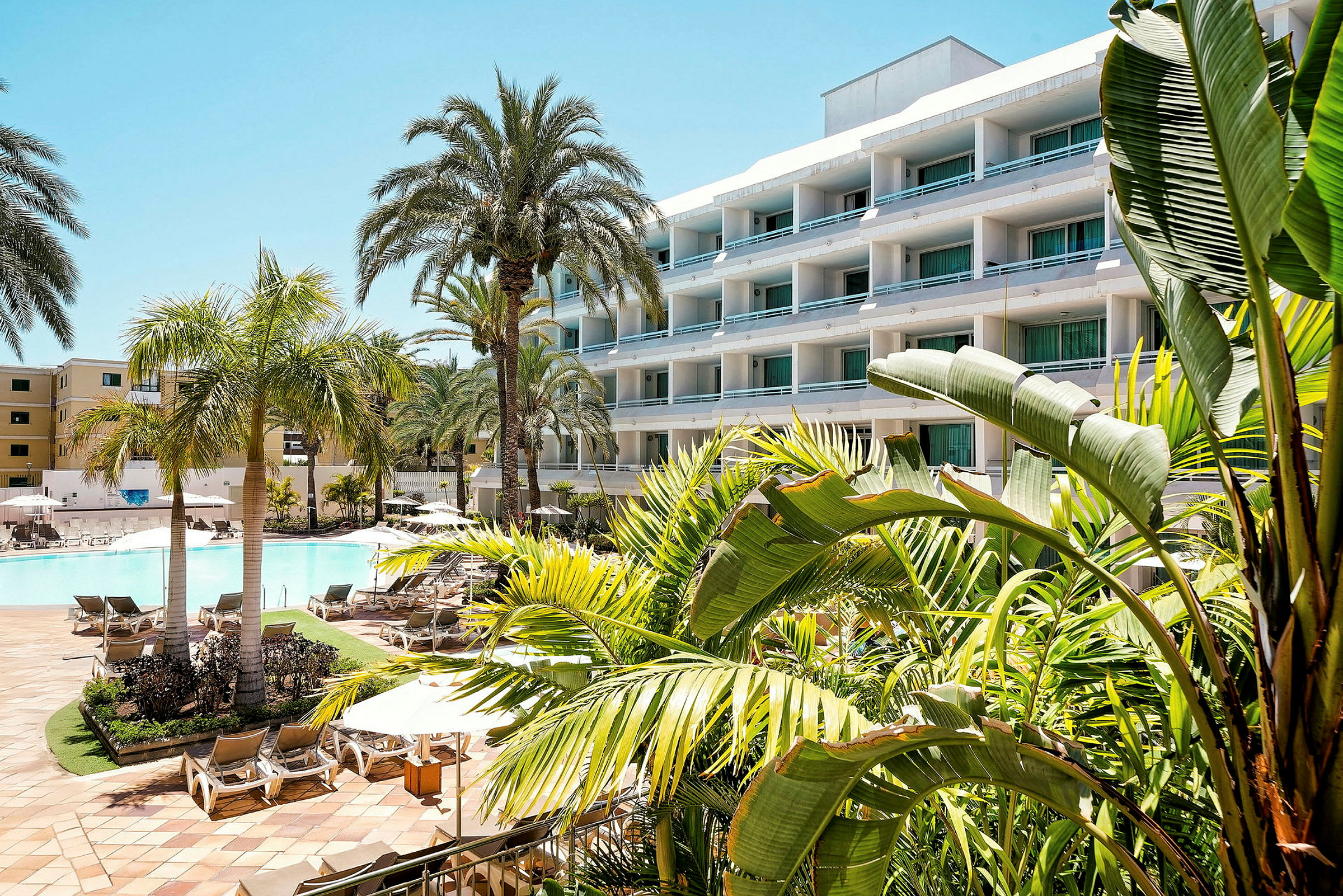 Hotellbyggnad med balkonger och utsikt över en pool, omgiven av palmer och grönska. (AI-genererad text)