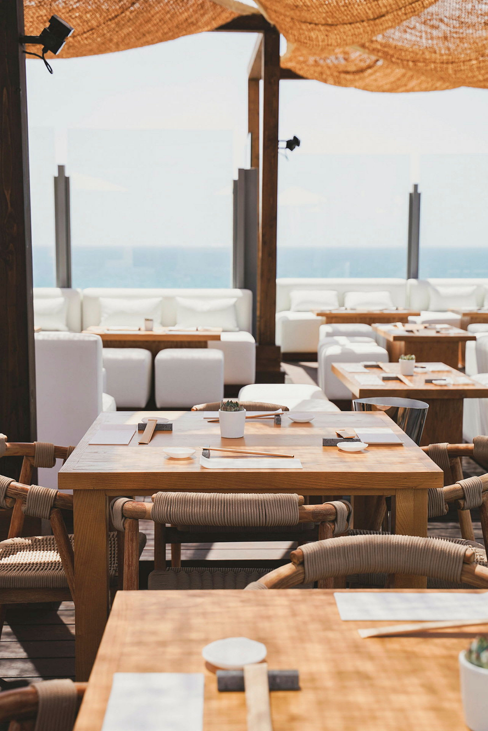 Utomhusrestaurang med träbord och stolar, vita soffor i bakgrunden, samt utsikt mot havet. (AI-genererad text)