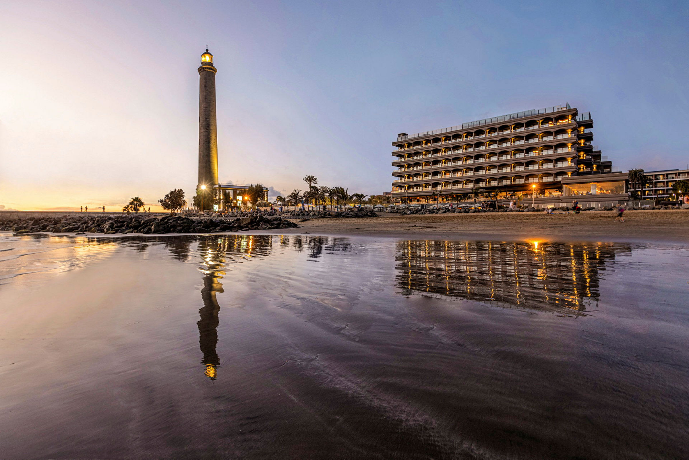 Bild av en fyr belägen vid stranden med ett hotell i bakgrunden, reflektioner synliga i vattenytan. (AI-genererad text)