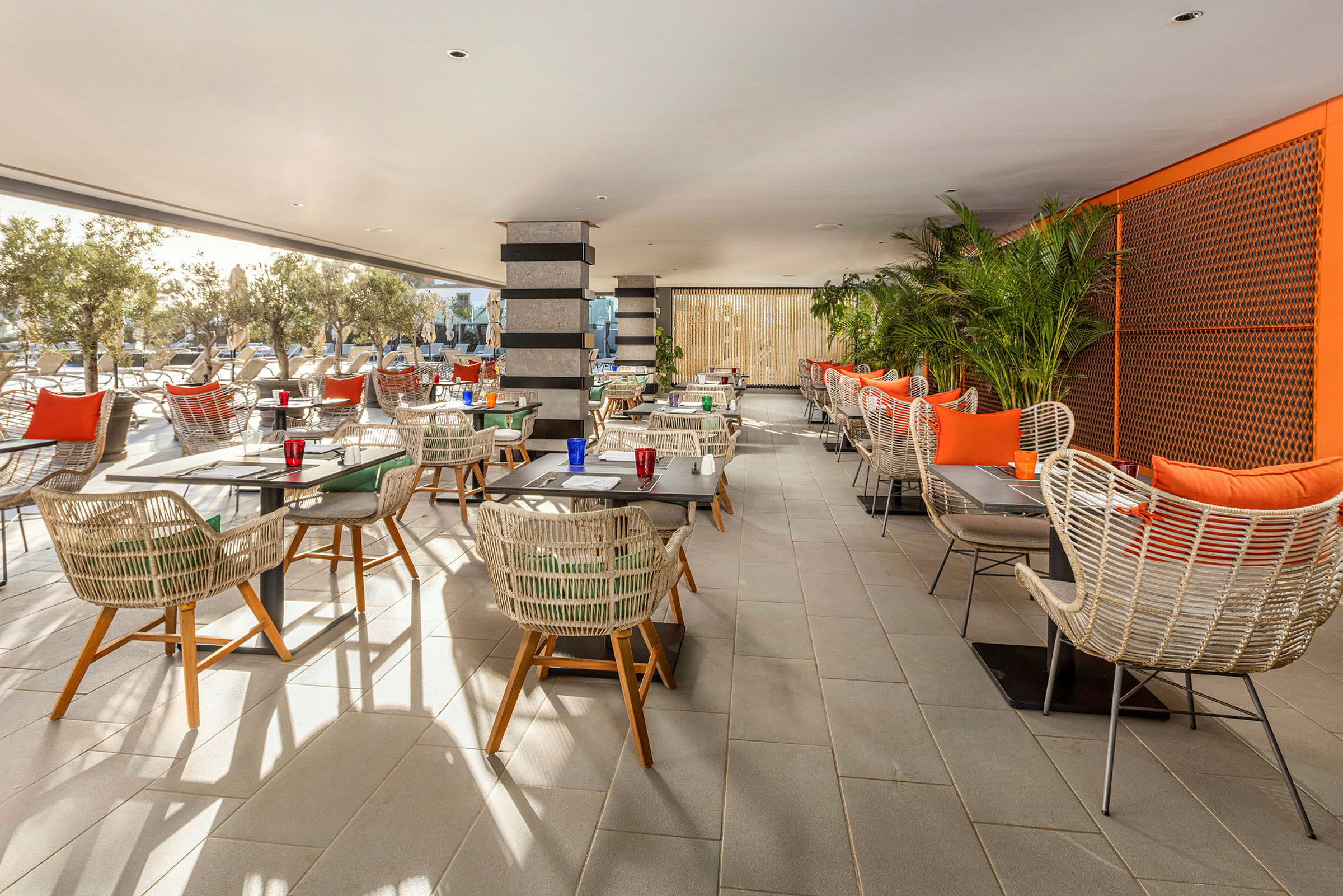 En restaurang med bord och stolar i rottingstil, omgiven av växter och utsikt mot en utomhusområde. (AI-genererad text)