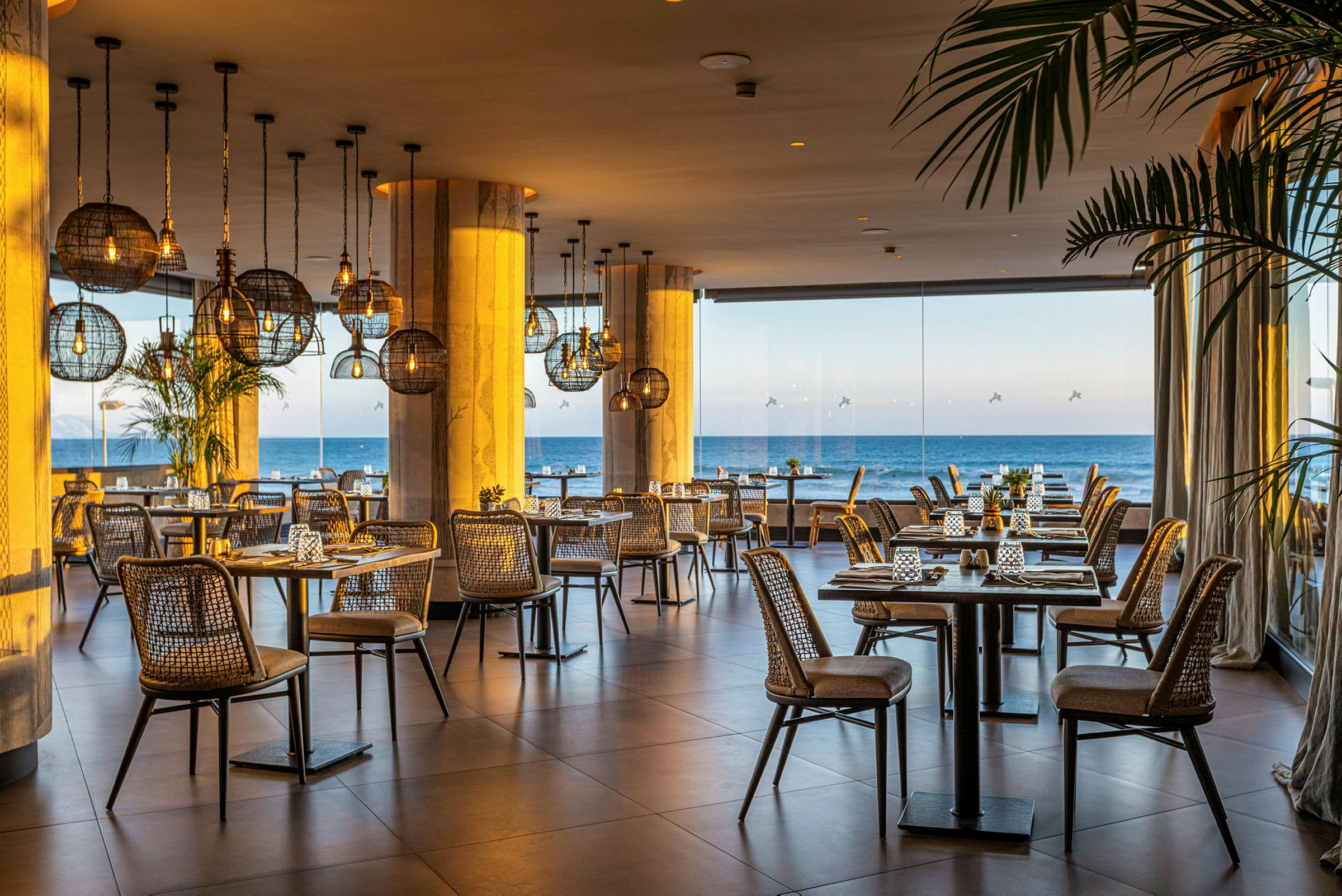 Bild av en restaurang med rottingstolar, borden är dukade med utsikt över havet genom stora glasfönster. (AI-genererad text)